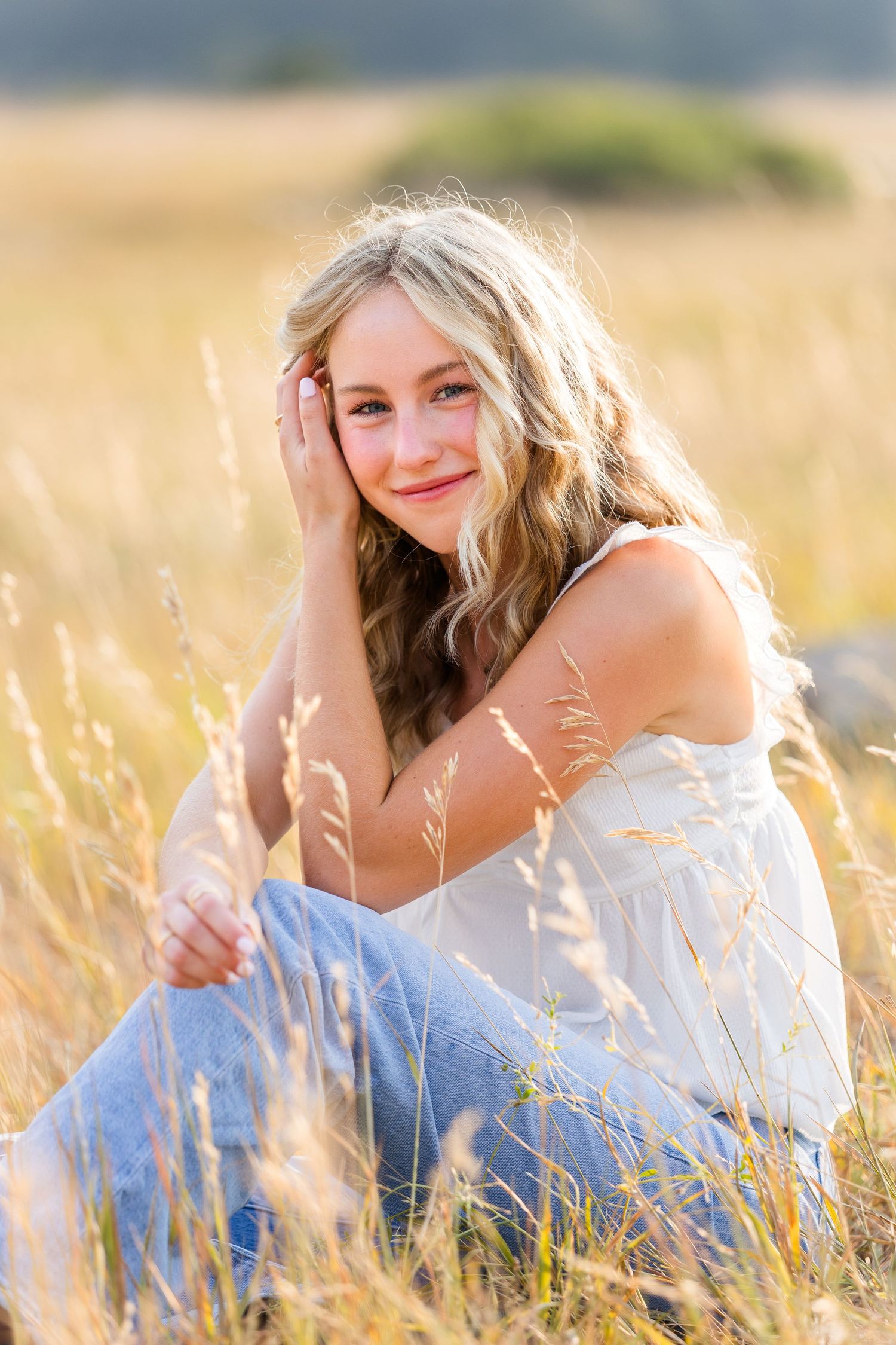 Smiling blonde teen girl sitting in golden field grass wearing white top and jeans in warm sunlight.