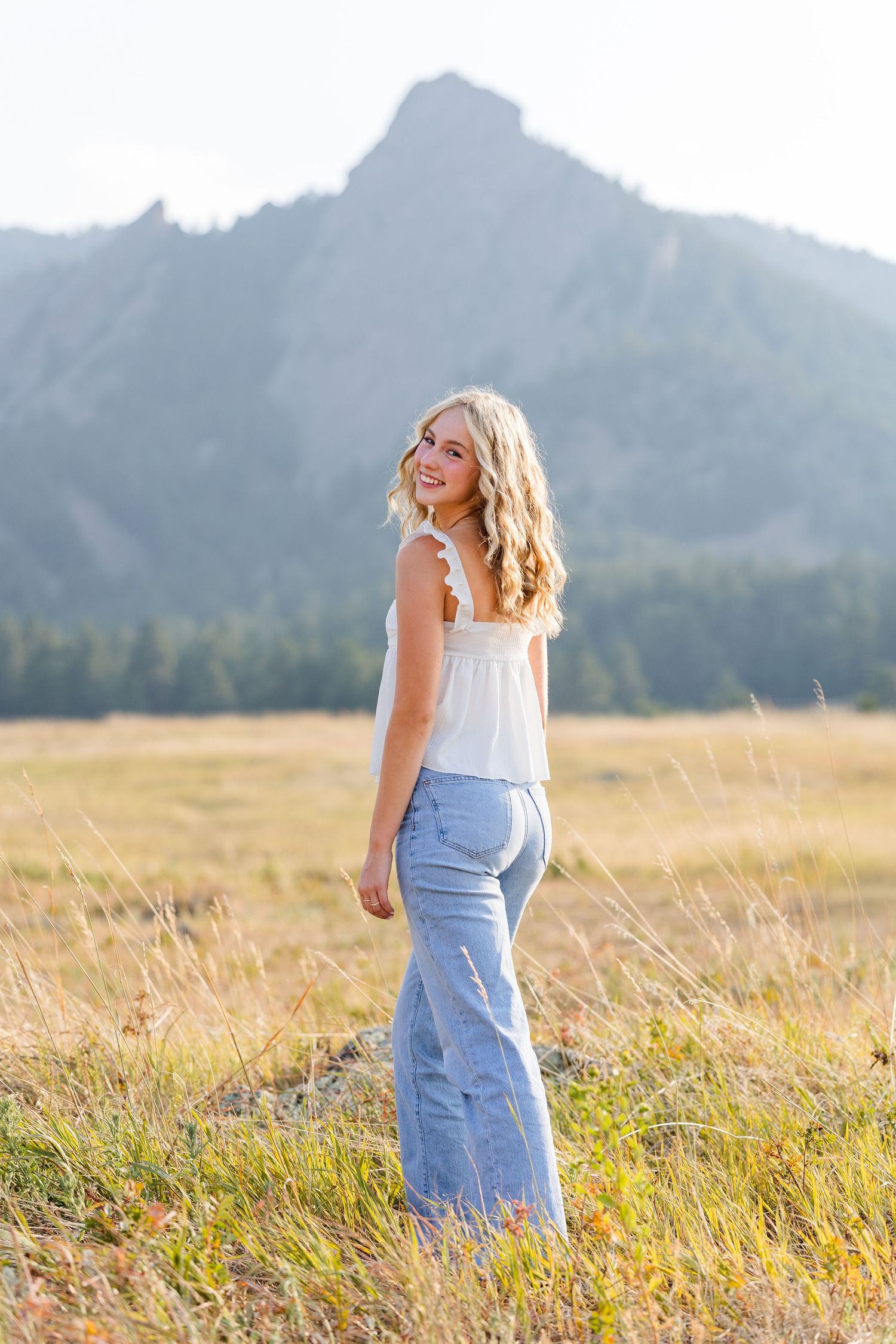 Smiling blonde teen girl in white top and flare jeans poses in golden meadow with rocky mountain peak behind her.