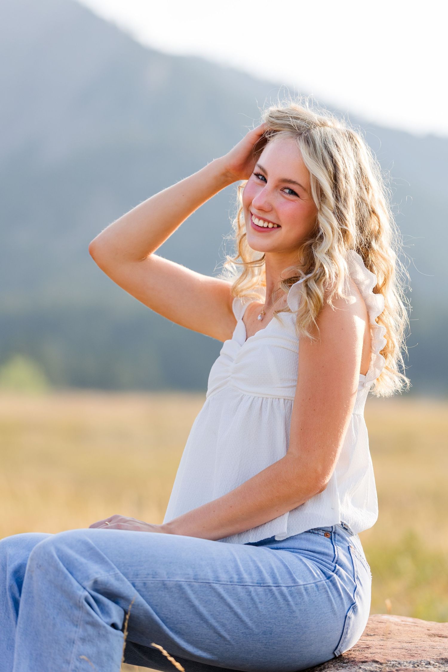 Smiling blonde woman in white top and jeans sits outdoors with mountains and golden field in background.