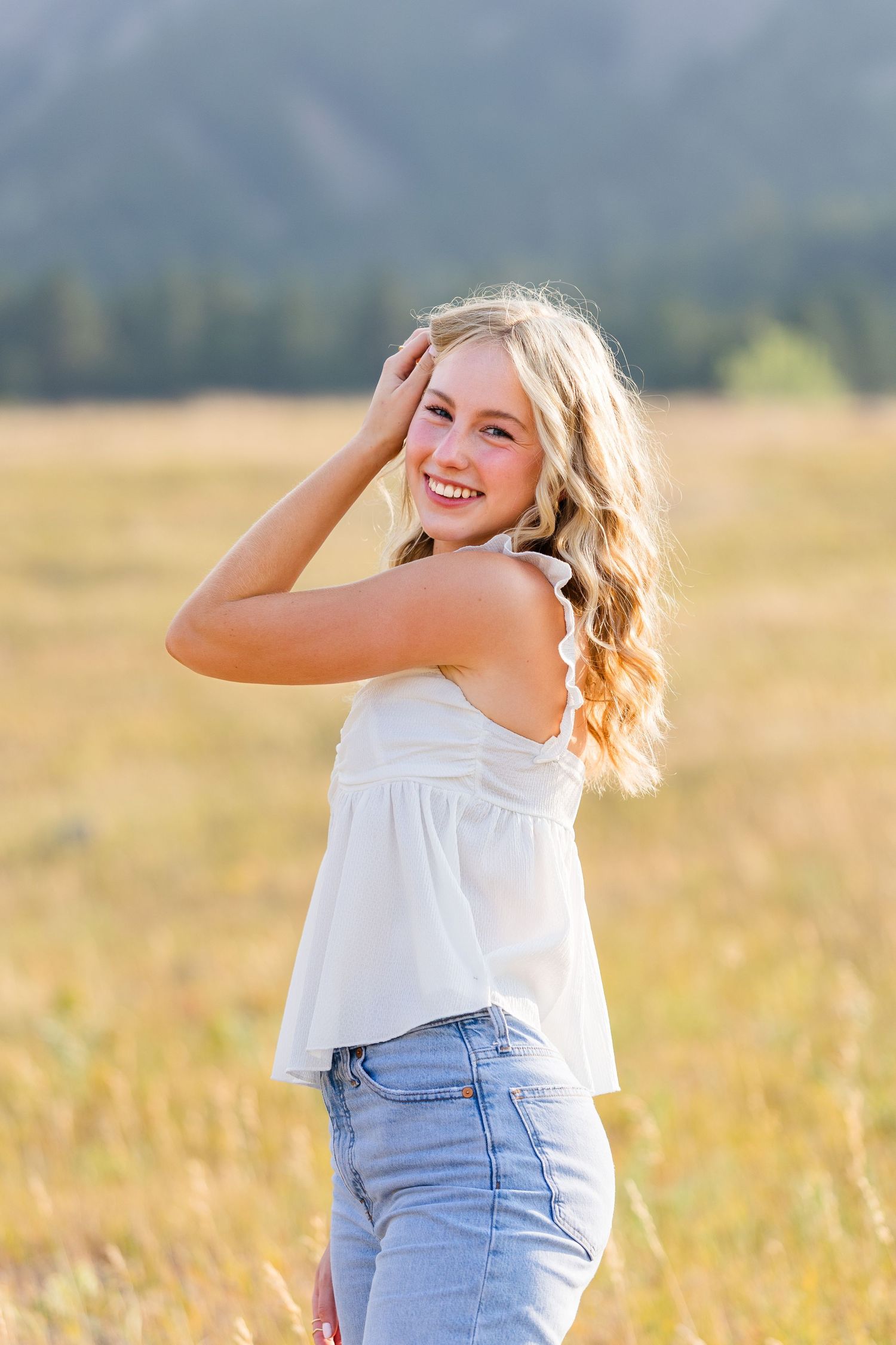 Smiling blonde teen girl in white top and jeans poses in golden field with mountains in background.