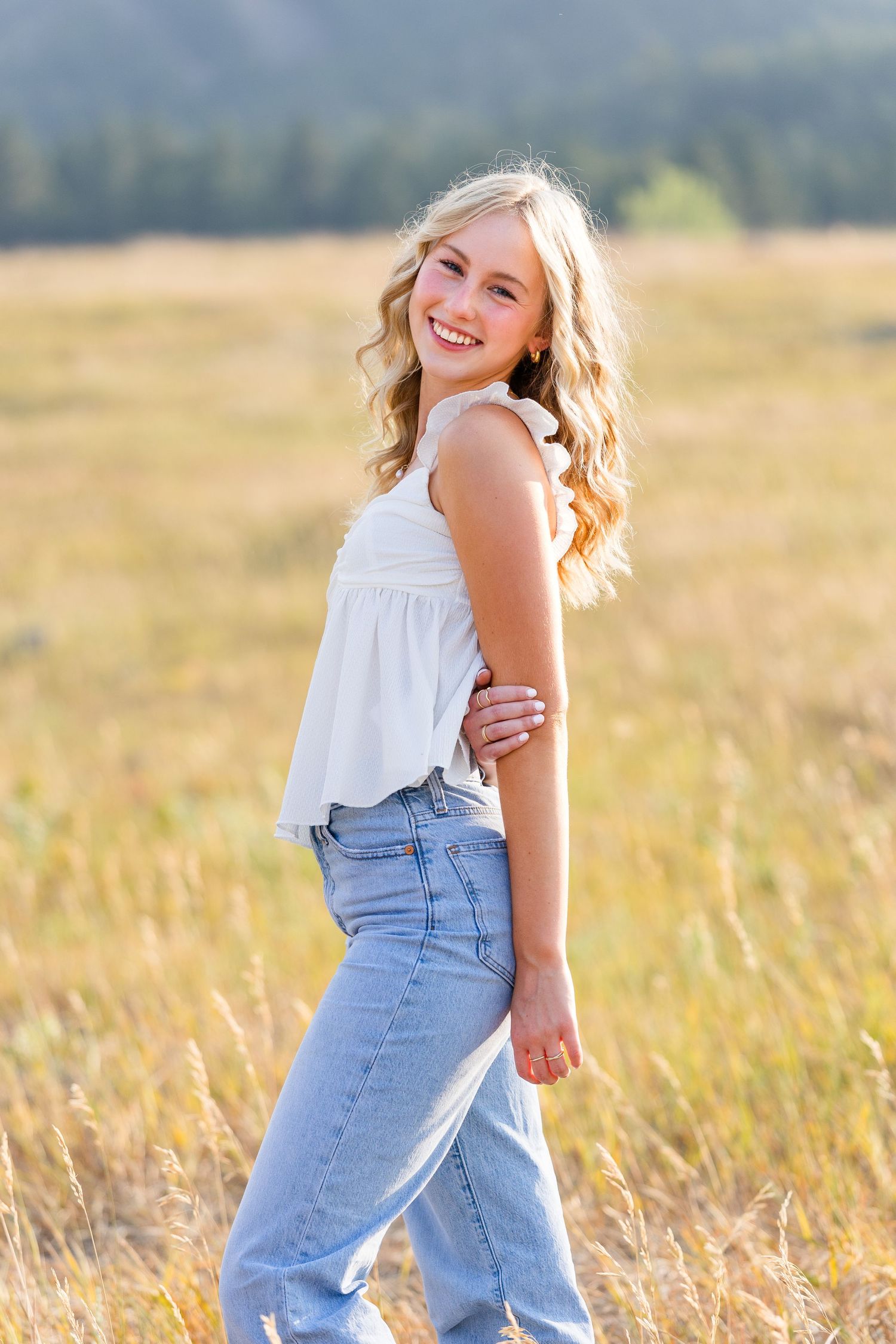 Happy blonde teenage girl in white top and jeans walks through sunlit golden field with mountains behind.
