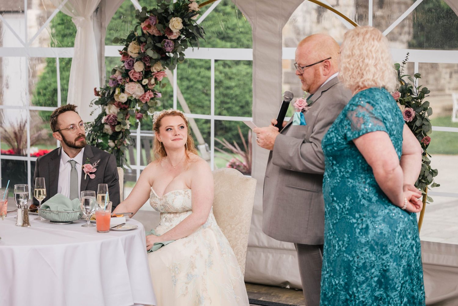 Bride's father reading from a note card during his toast, bride watching with a soft smile.