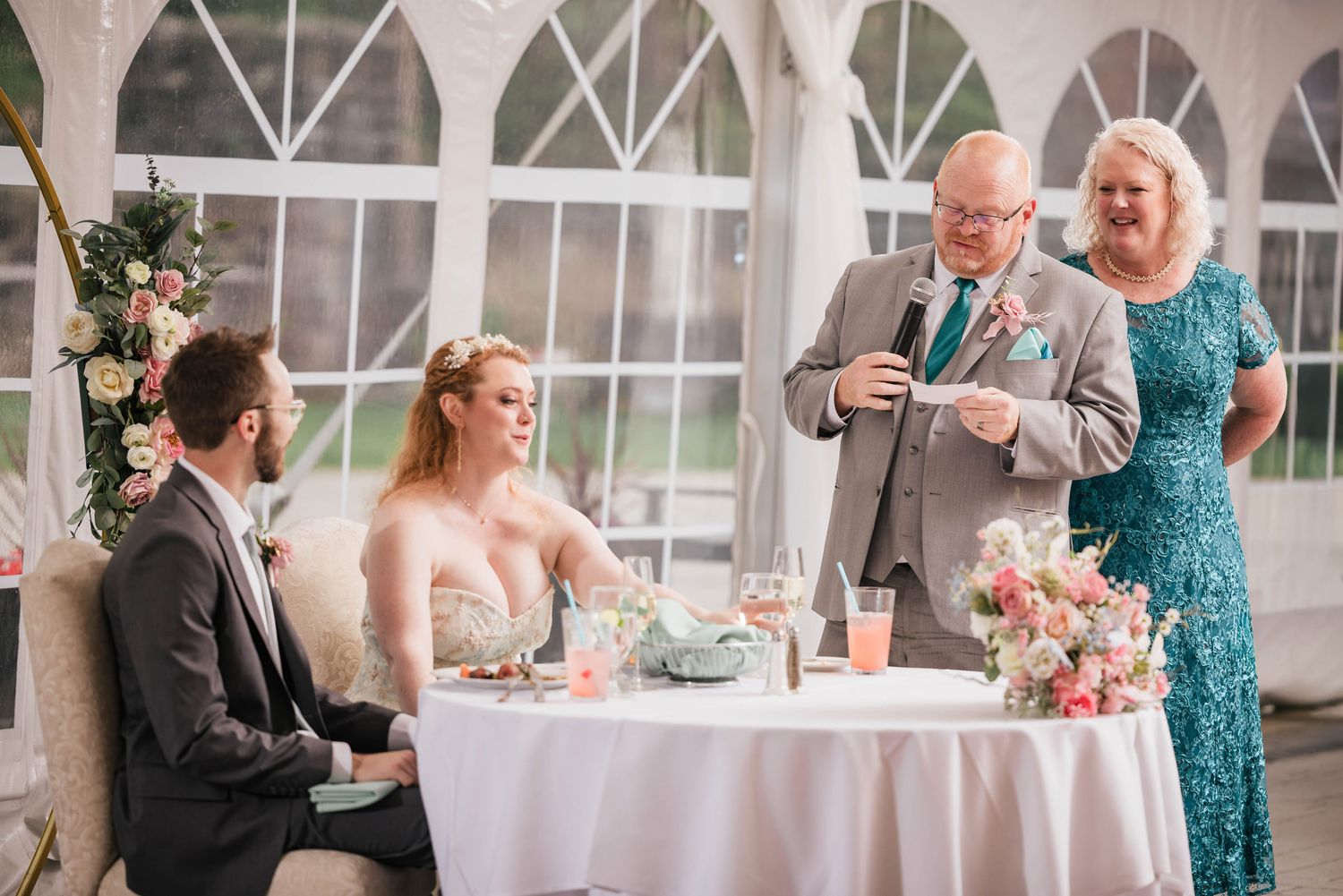 Bride's father giving a speech at the sweetheart table, bride's mother standing beside him laughing.