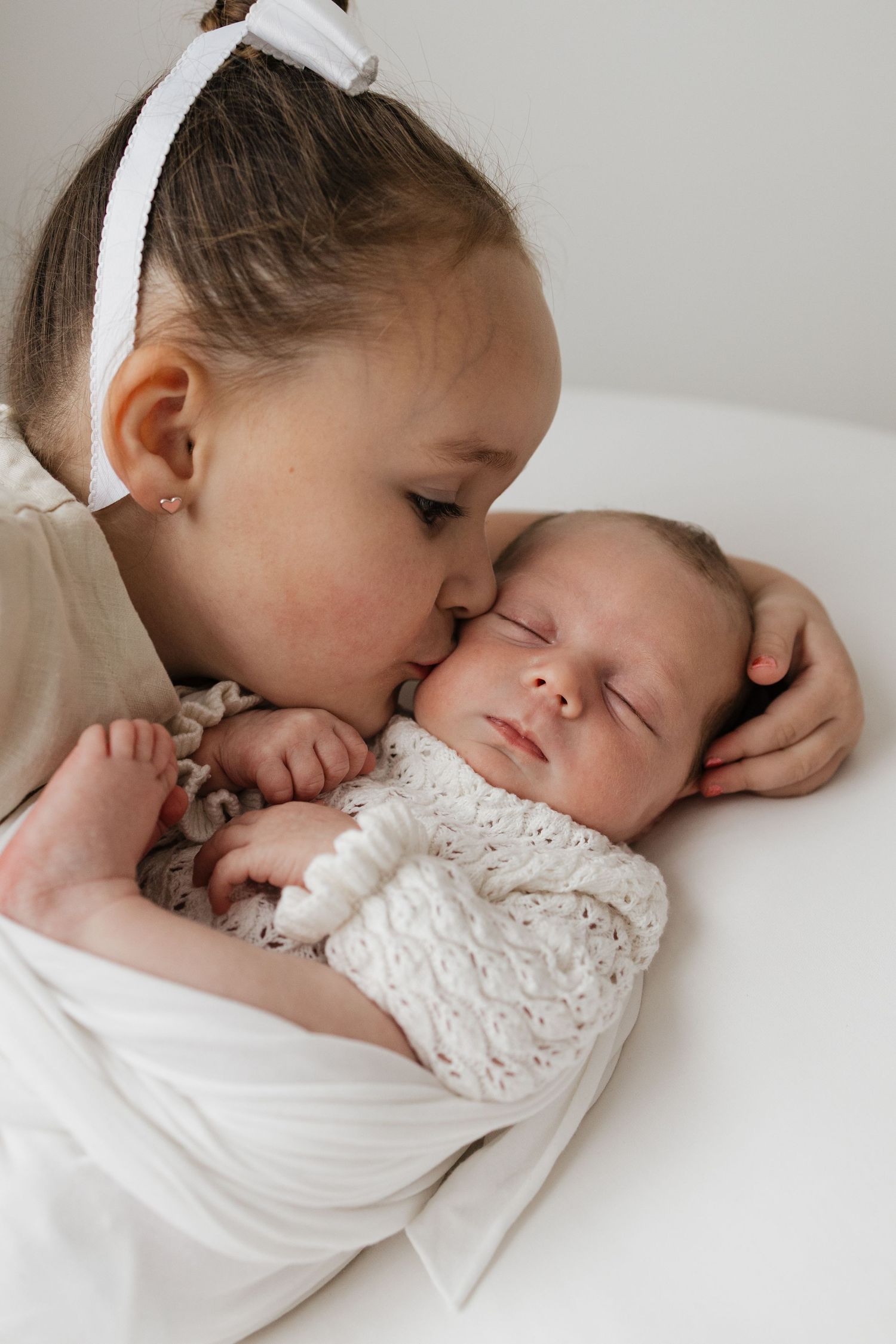 Two young girls sitting on a beige couch smiling while gently holding a sleeping newborn baby between them.