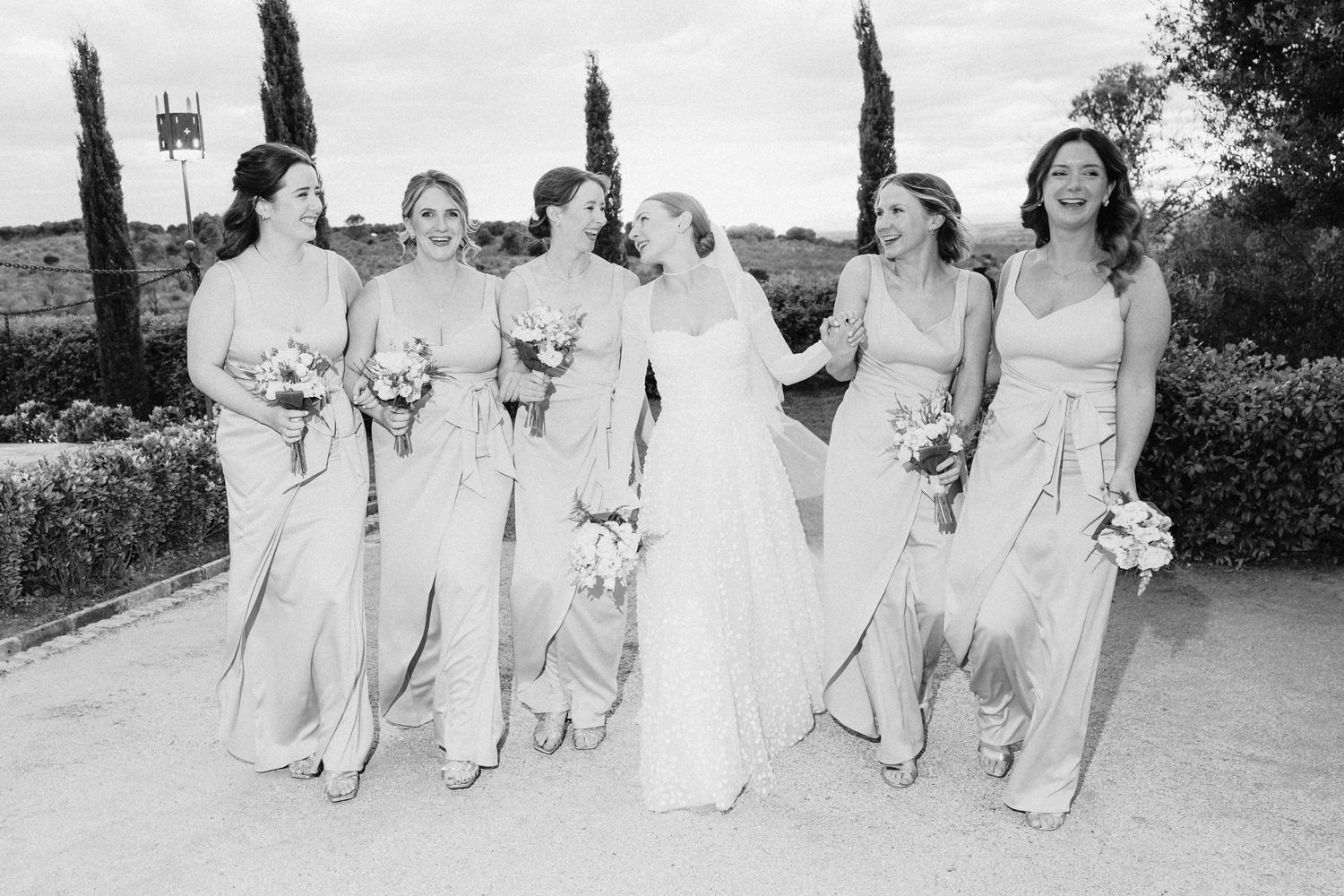 Bride walks joyfully with five bridesmaids in matching gowns holding bouquets in a black and white outdoor photo.