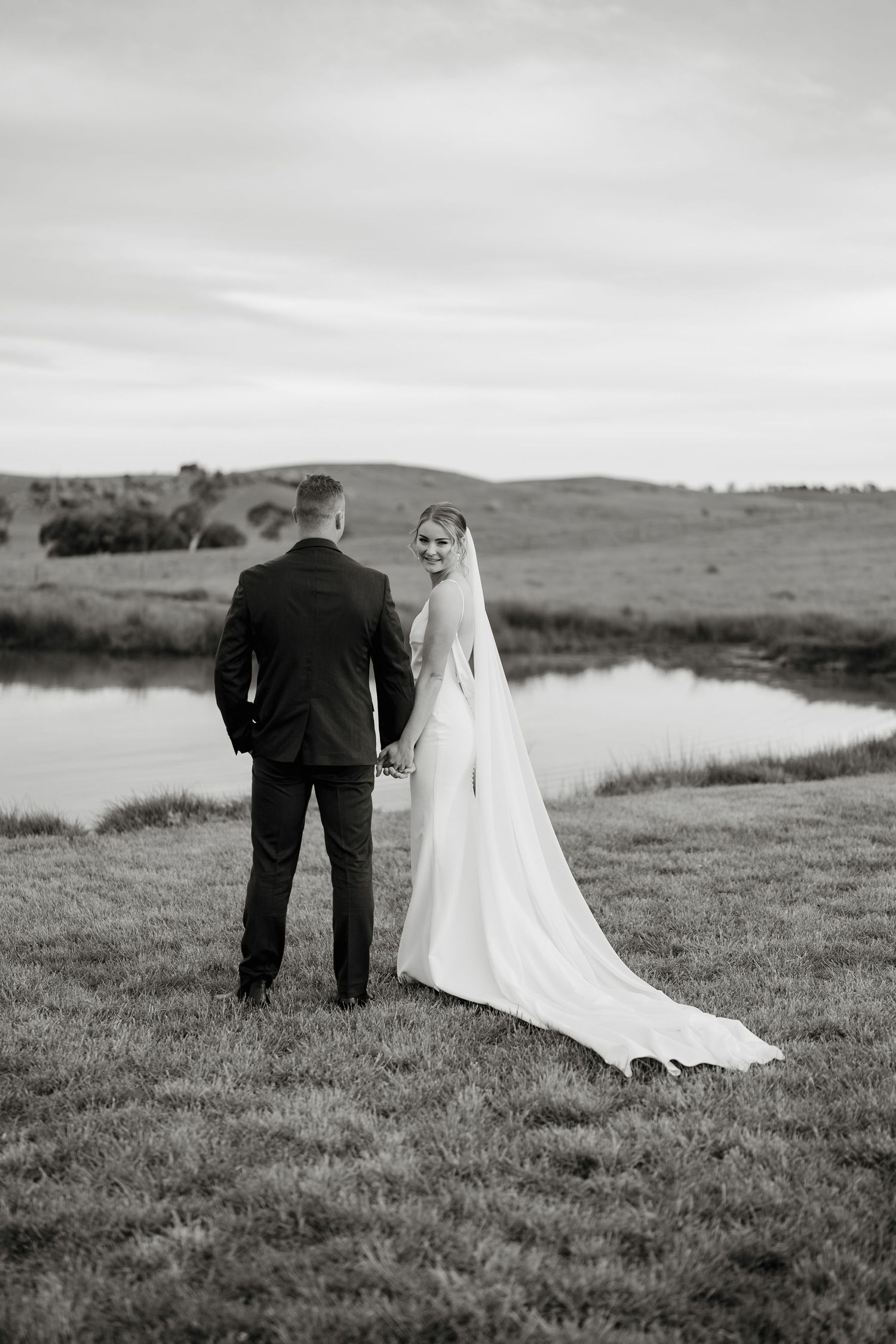 Black and white photo of bride and groom holding hands by a serene pond, bride glancing back with flowing veil and train. The Hayshed Orange