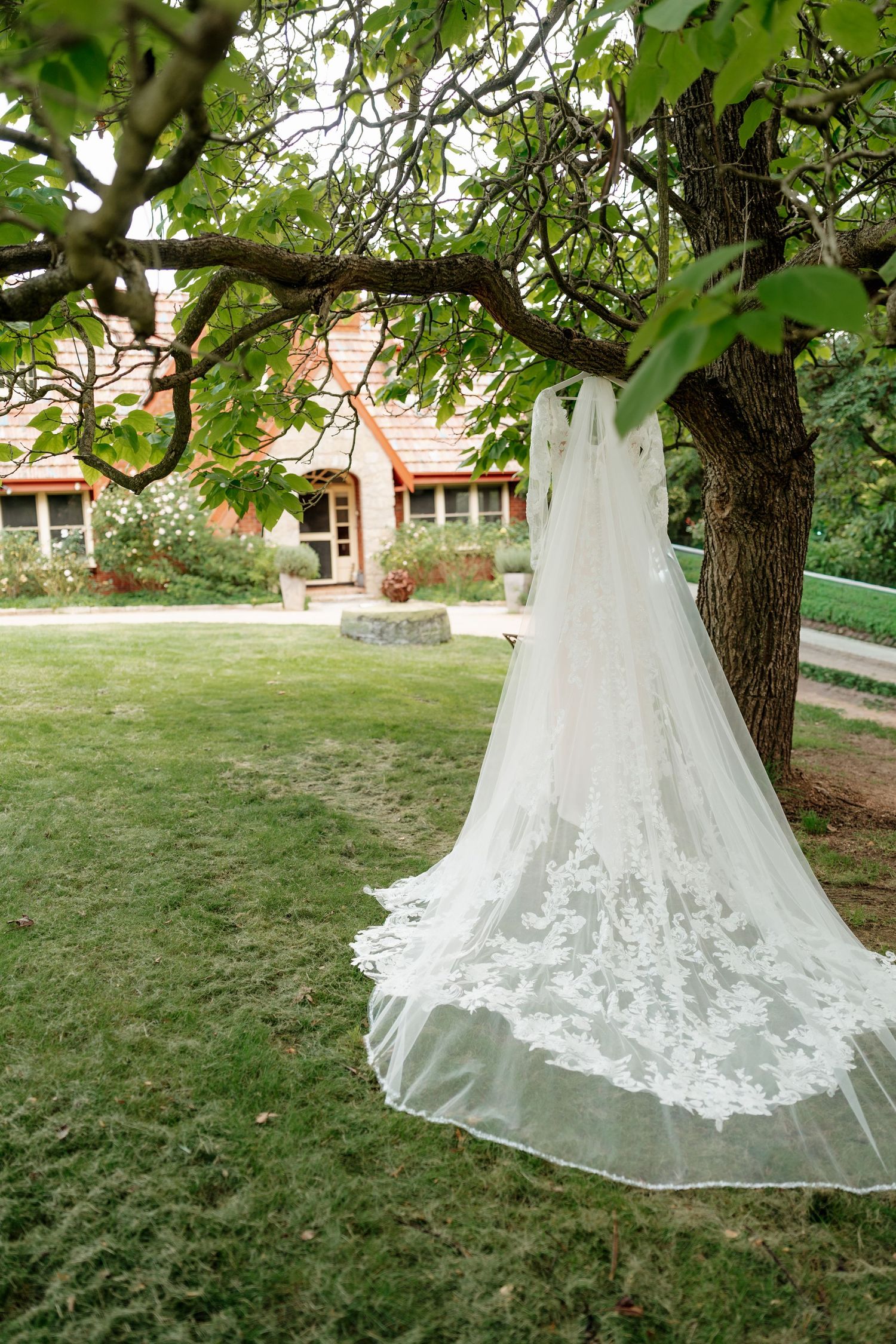Elegant lace wedding dress hanging from a tree branch in a lush garden with a charming house in the background. Clifton Woolshed Bathurst.