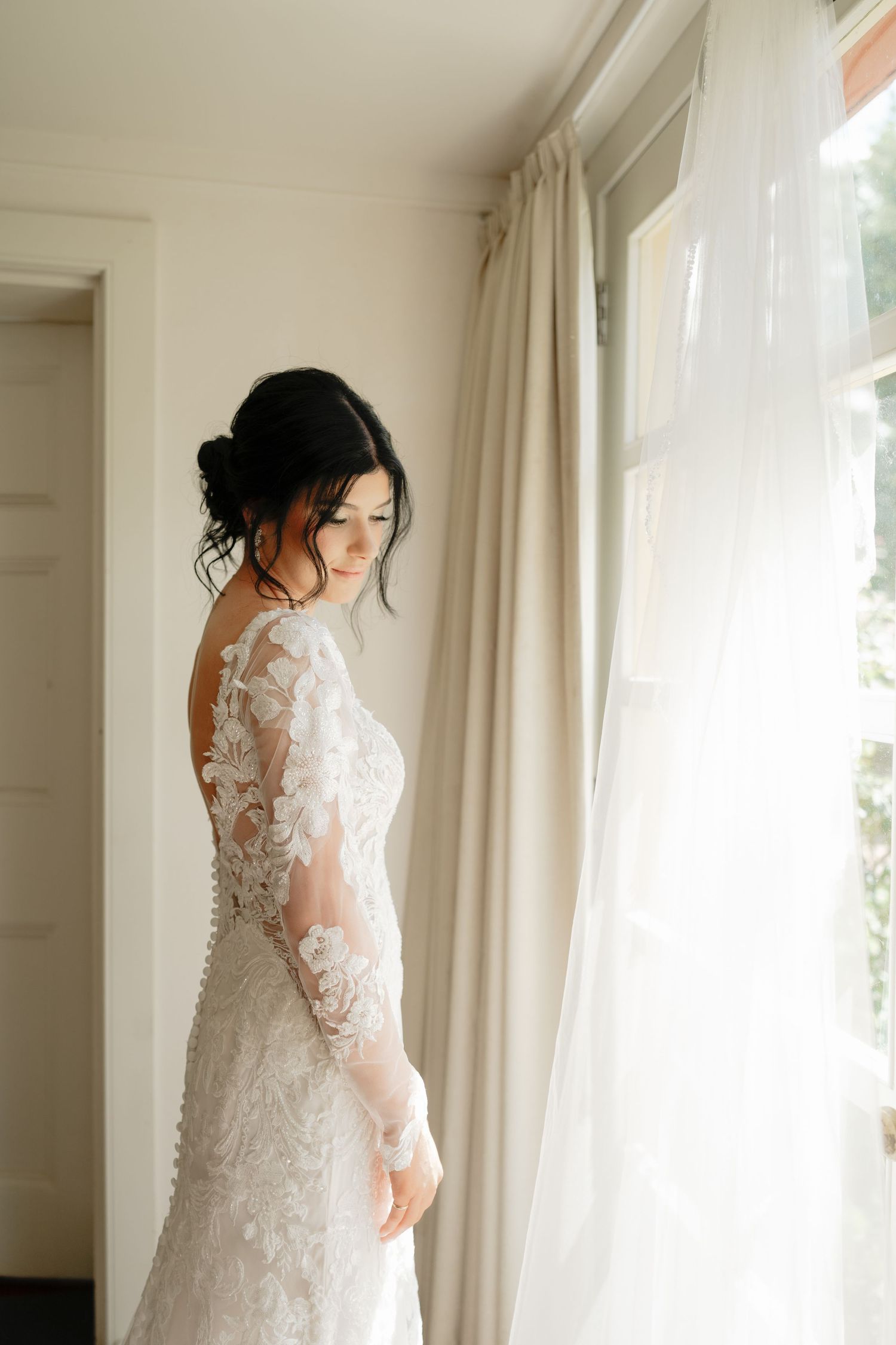 Bride in lace long-sleeve wedding gown standing by a sunlit window, looking down gracefully. Clifton Woolshed Bathurst.