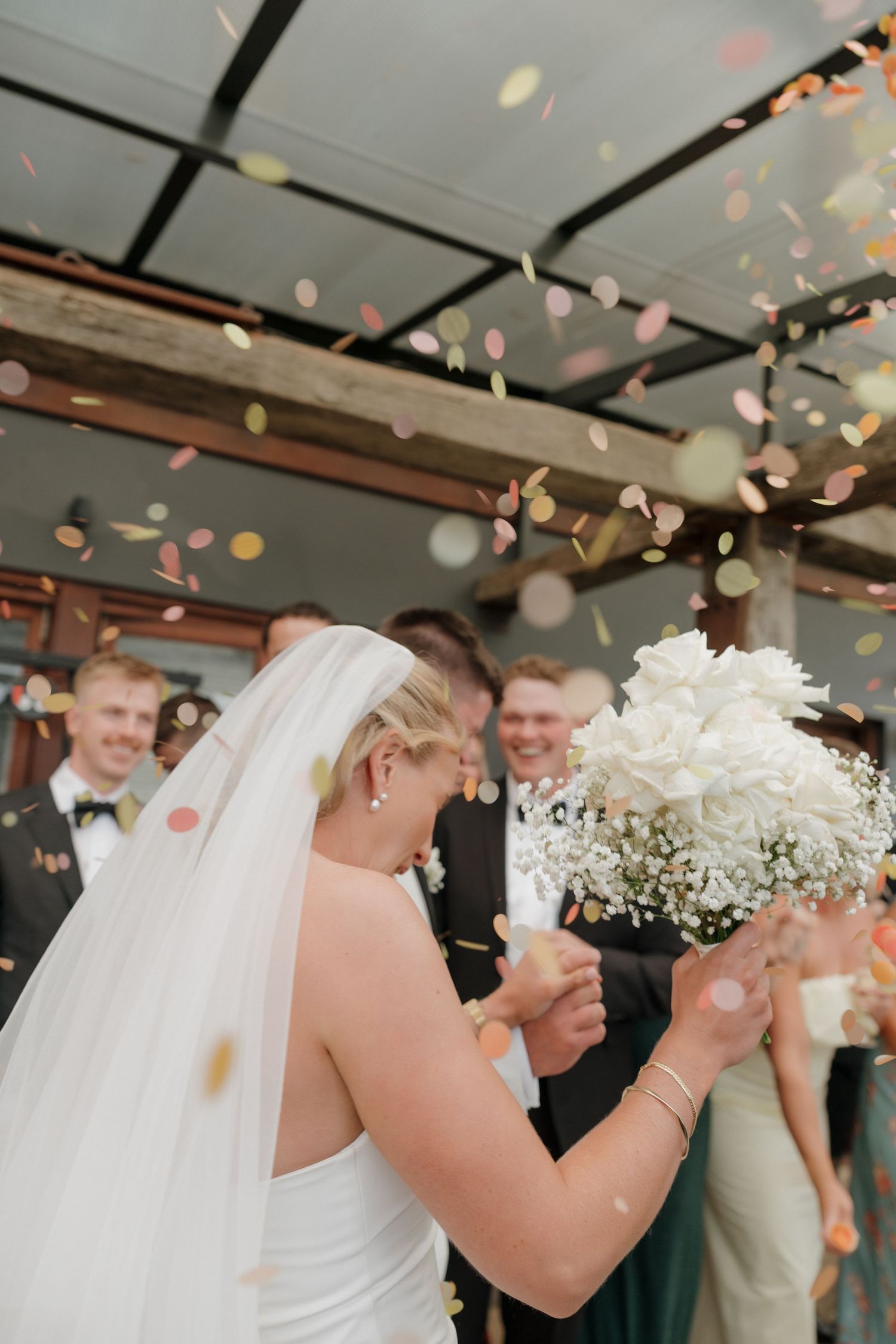 Bride in white gown and veil holds bouquet as colorful confetti falls during outdoor wedding celebration. Borrodell Vineyard Orange NSW