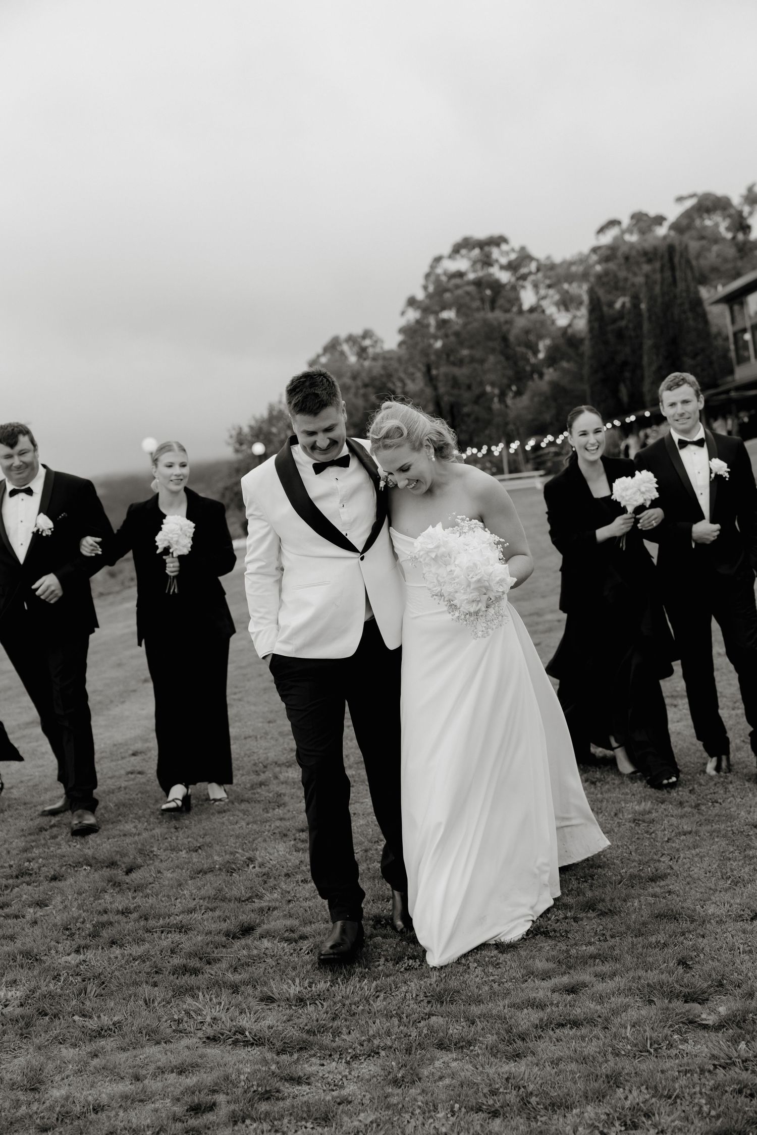 Black and white photo of bride and groom walking with wedding party on grass at outdoor ceremony. Borrodell Vineyard Orange.