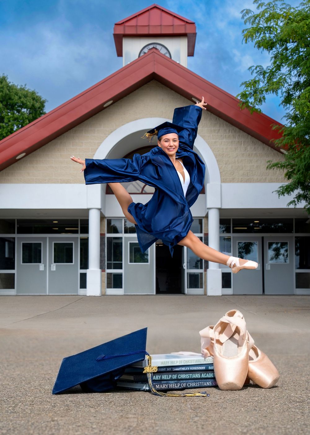 Graduate in blue cap and gown leaps gracefully in ballet pose with pointe shoes and books on ground.