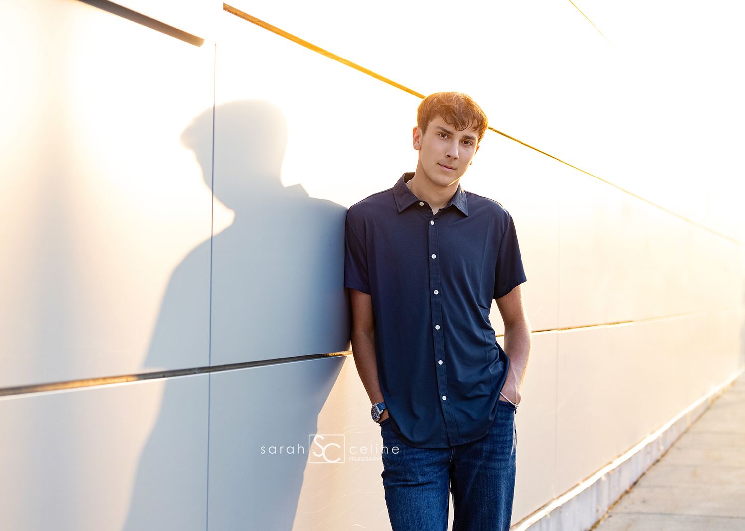A young man in a navy shirt and jeans leans against a modern wall, casting a dramatic shadow in warm light.