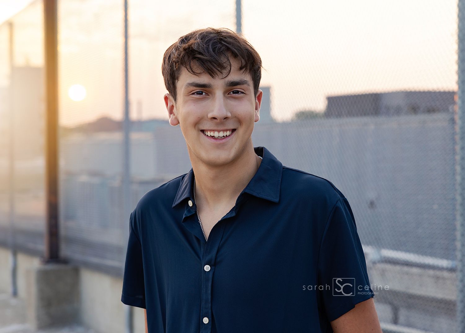 Smiling young man in navy blue polo shirt poses outdoors at golden hour with urban background.