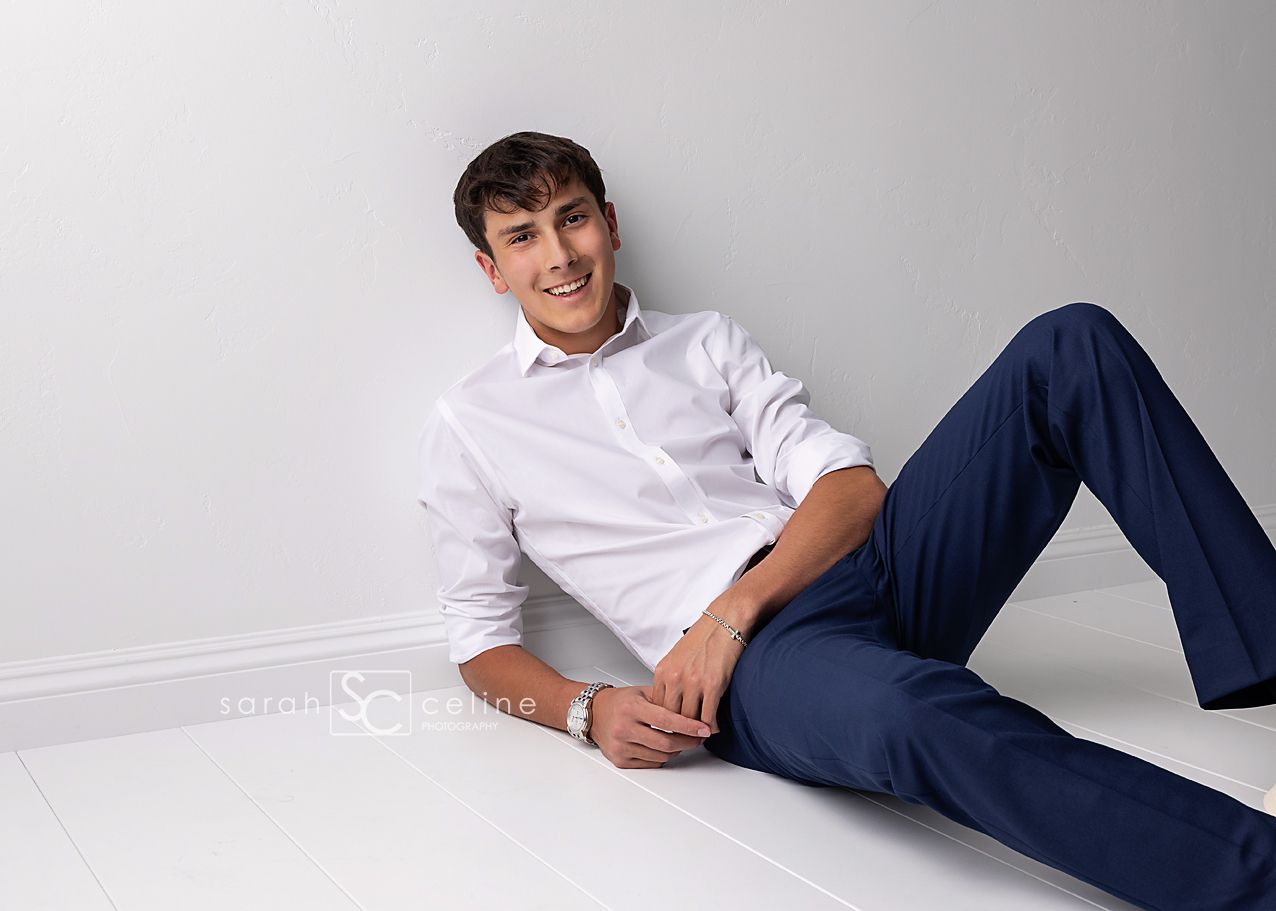 Young man in white shirt and navy pants poses casually on white floor against gray wall, smiling confidently.