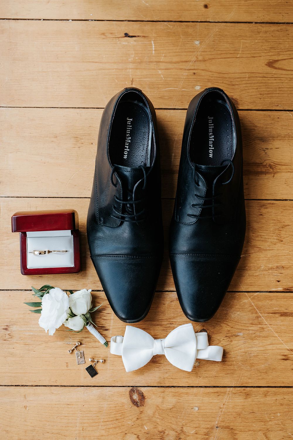 Black dress shoes, white bow tie, wedding rings in red box, and white boutonniere arranged on wooden floor.