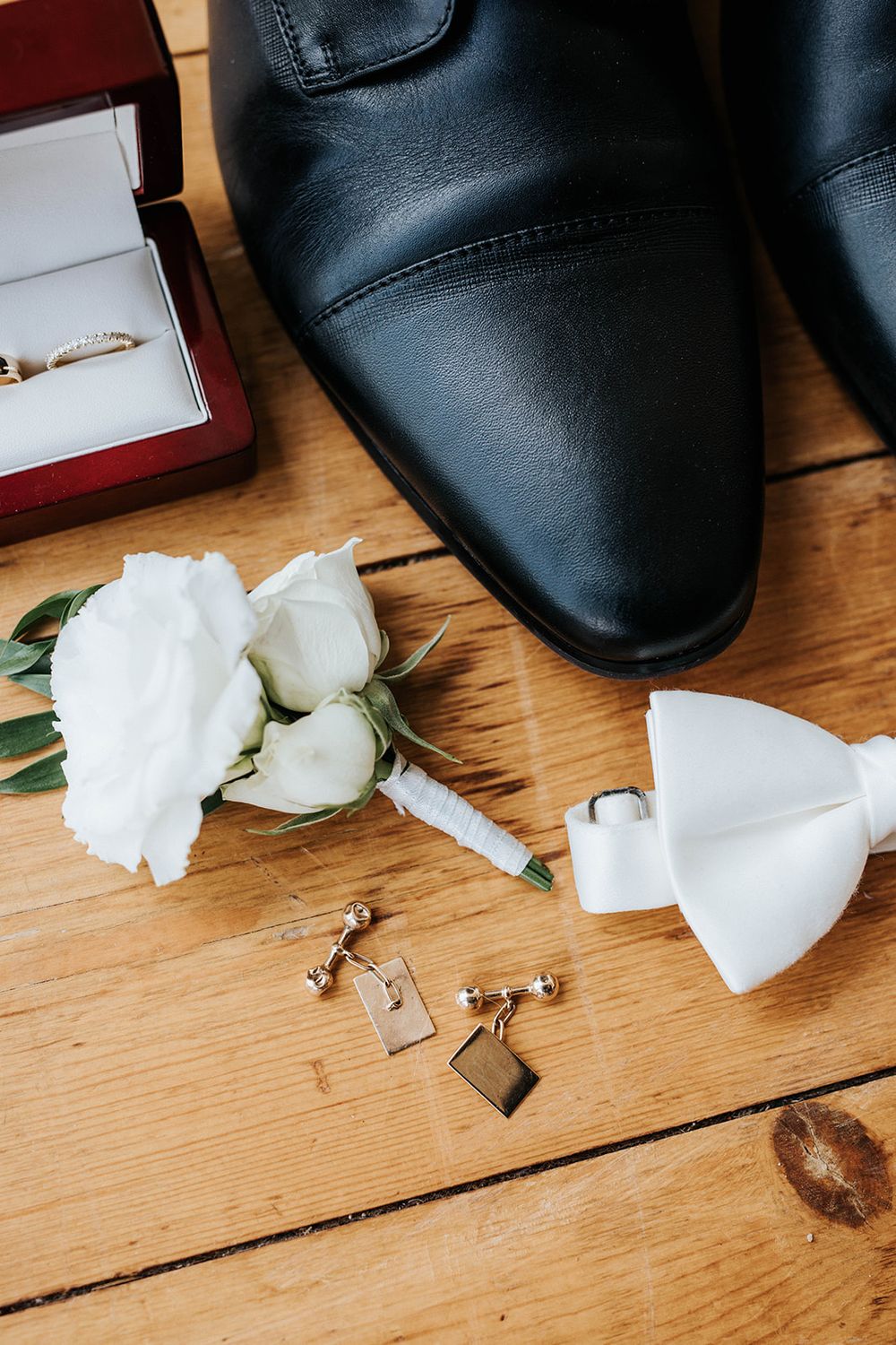 Wedding flat lay with black leather shoes, white boutonniere, bow tie, gold cufflinks, and ring box on wooden floor.