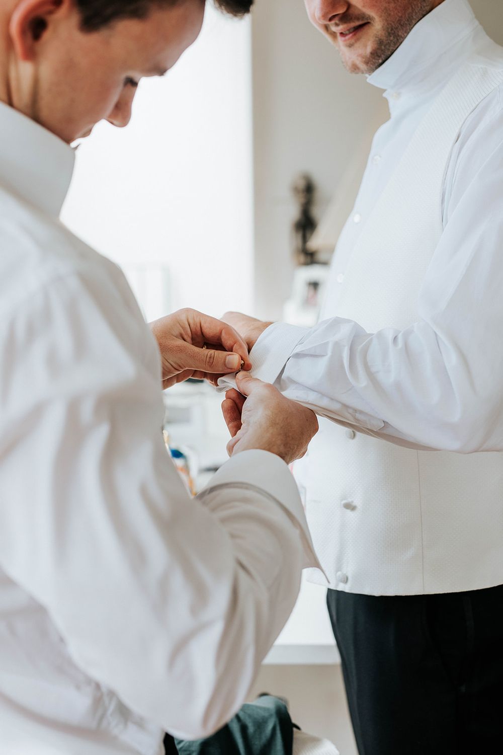Two grooms in white dress shirts helping each other fasten cufflinks while smiling on wedding day.