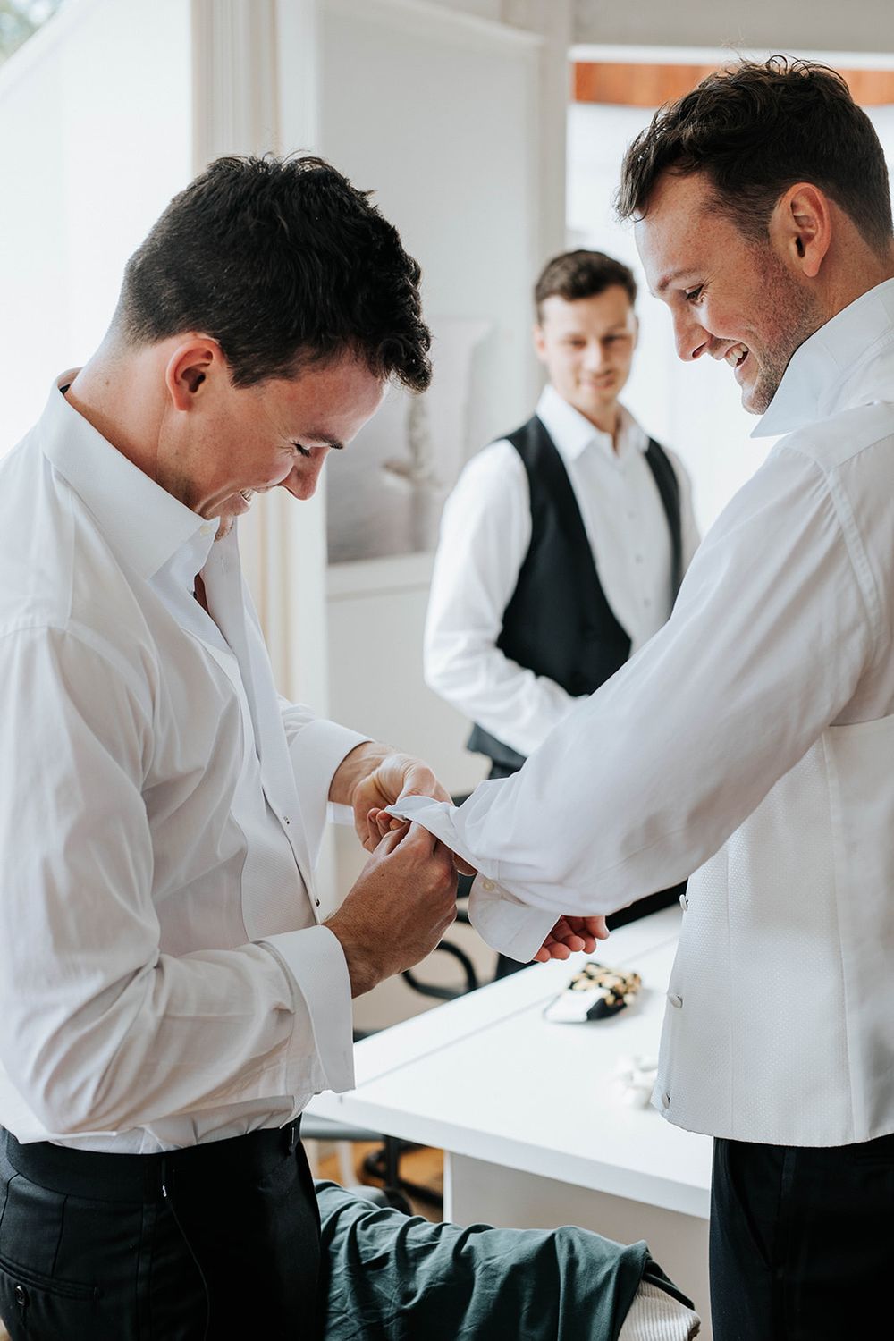 Two groomsmen in white shirts help each other with cufflinks while a third man in a vest watches during wedding preparations.