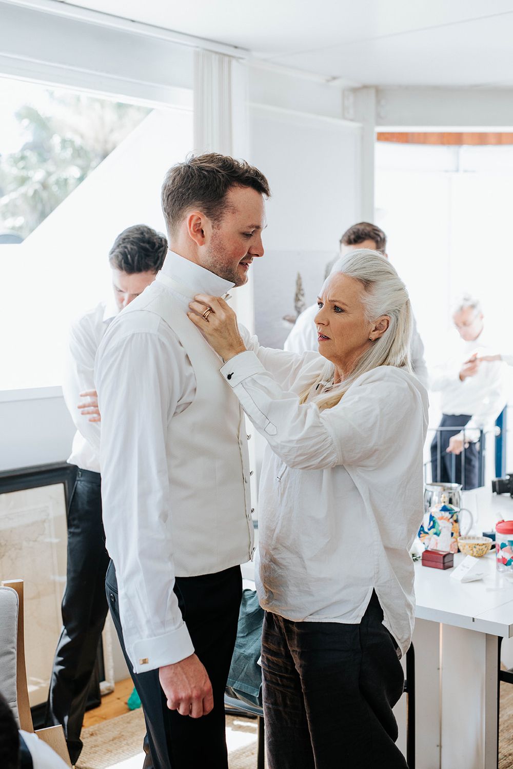 A woman helps a groom button his white dress shirt while getting ready for a wedding ceremony indoors.