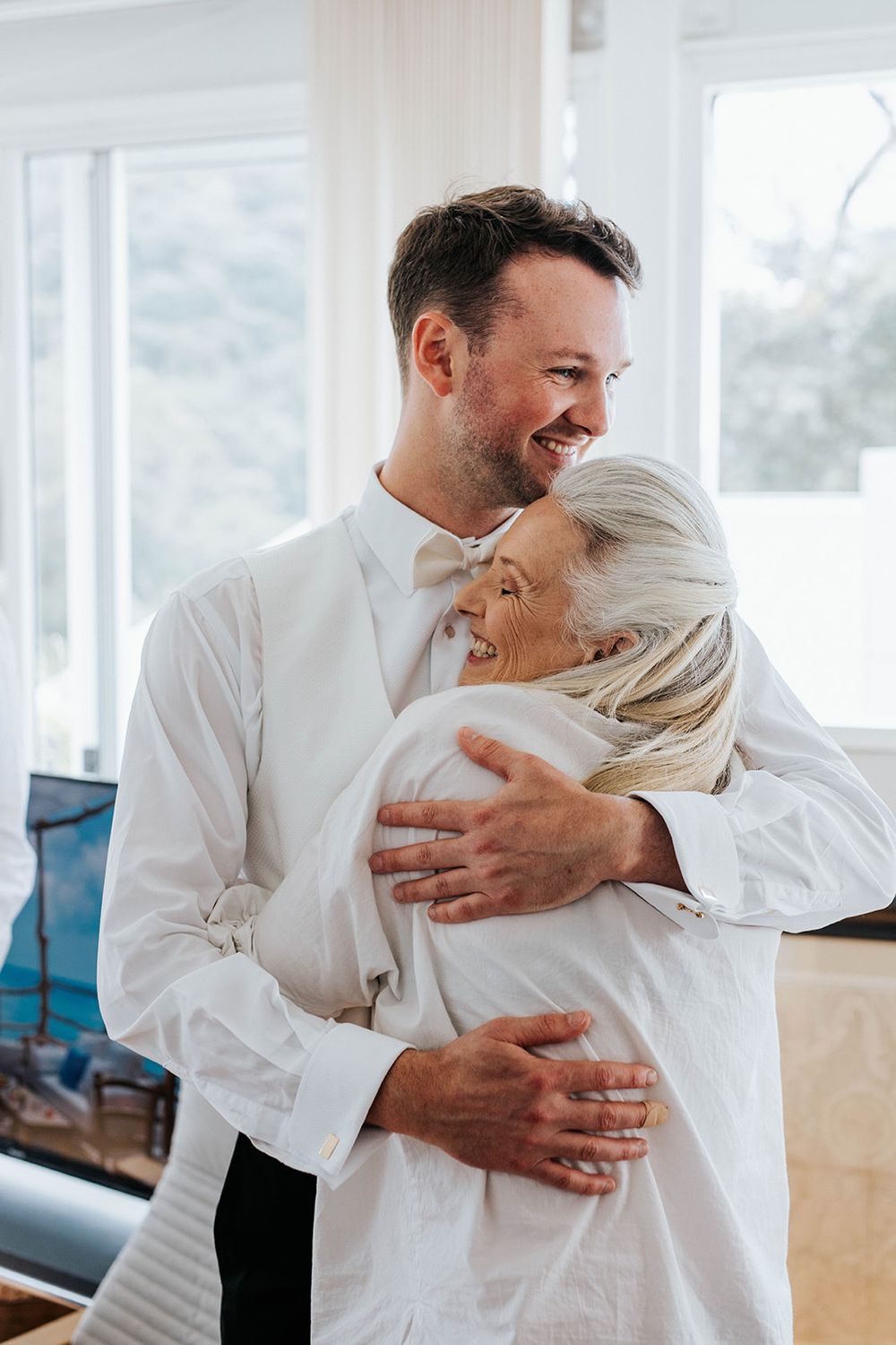 A smiling groom in a white suit hugs an older woman with silver hair during a joyful wedding celebration indoors.