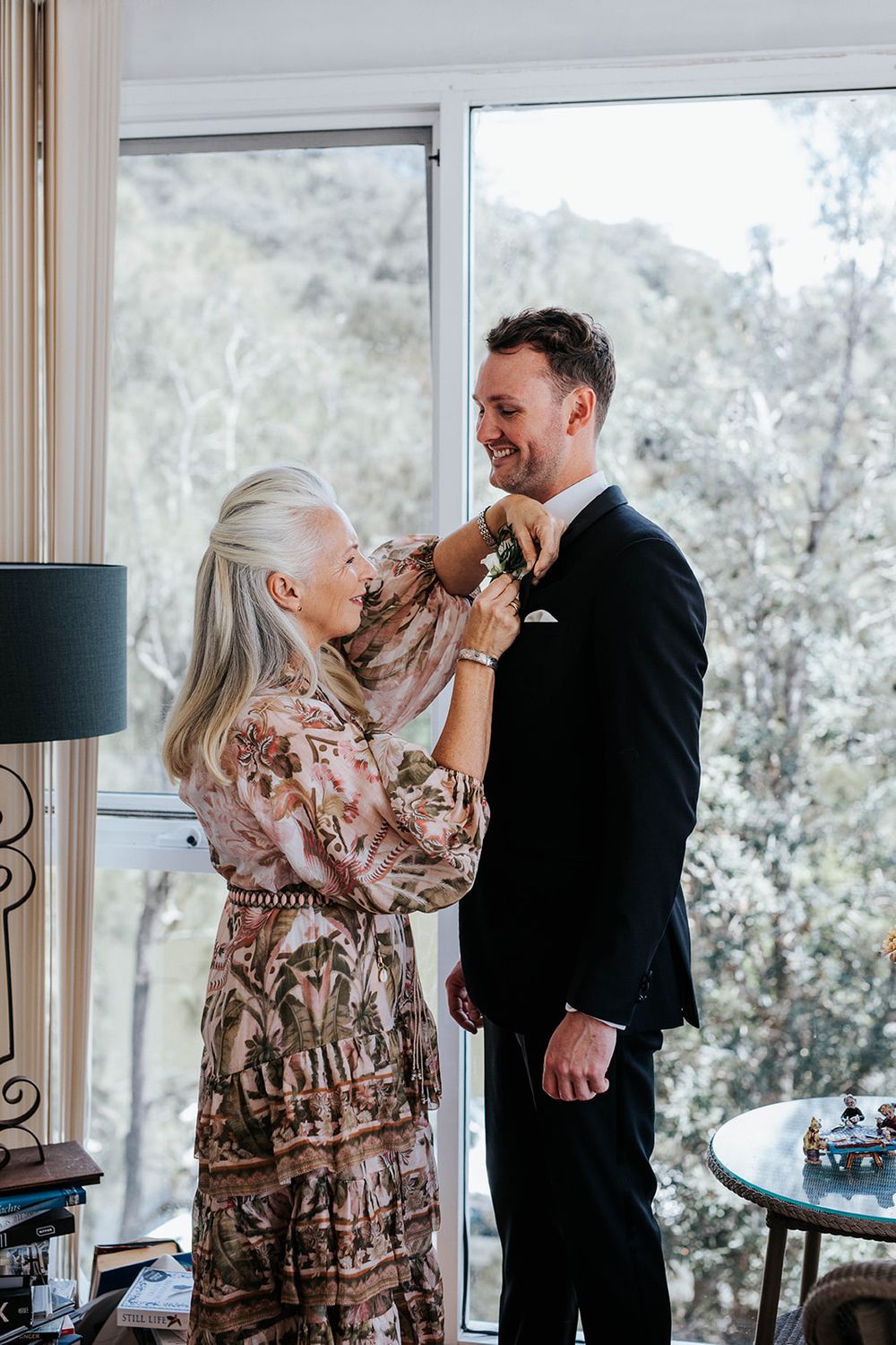A woman in a floral dress pins a boutonniere on a smiling groom in a black suit near a bright window.