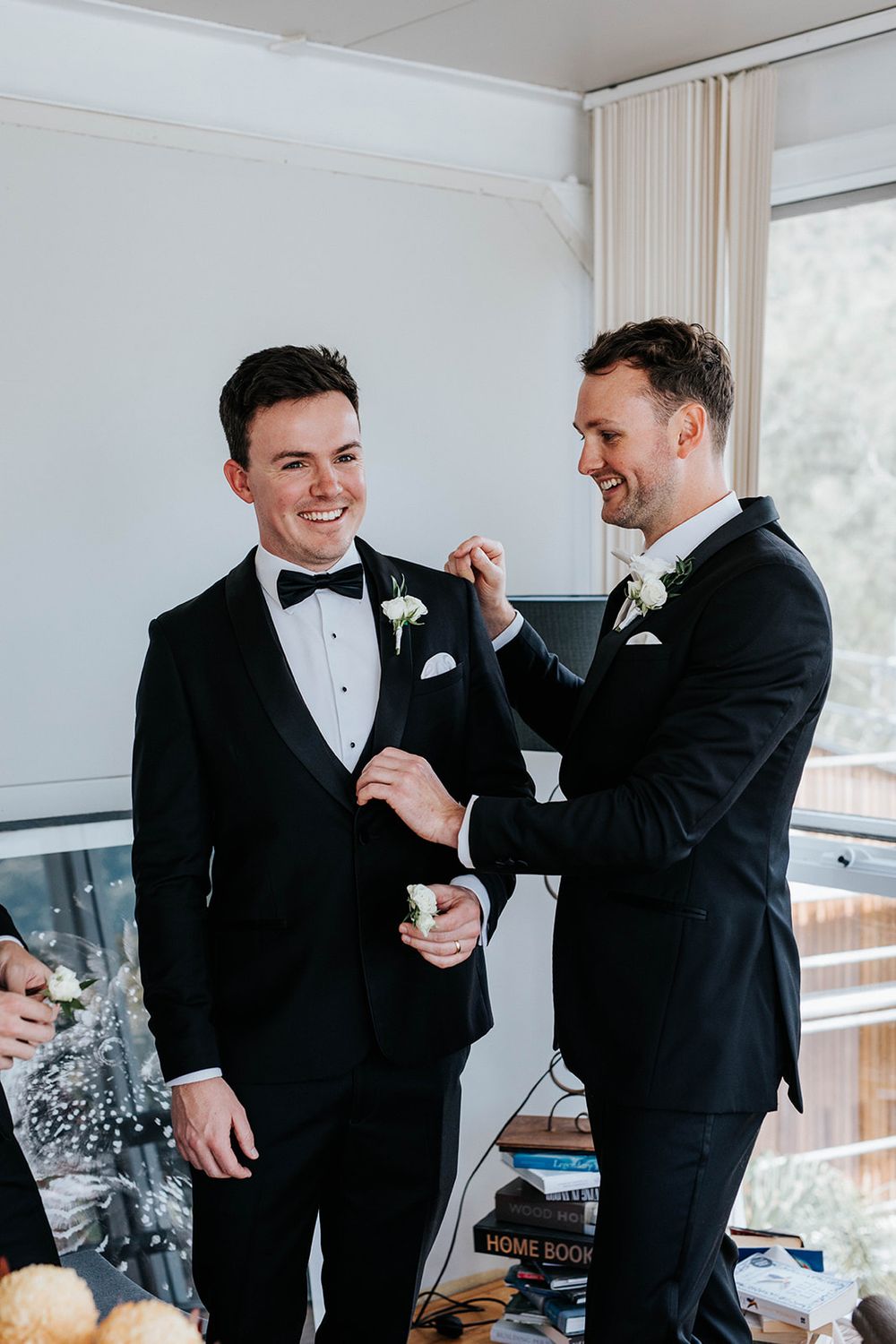 Two groomsmen in black tuxedos laughing as one adjusts the other's boutonniere before wedding ceremony.