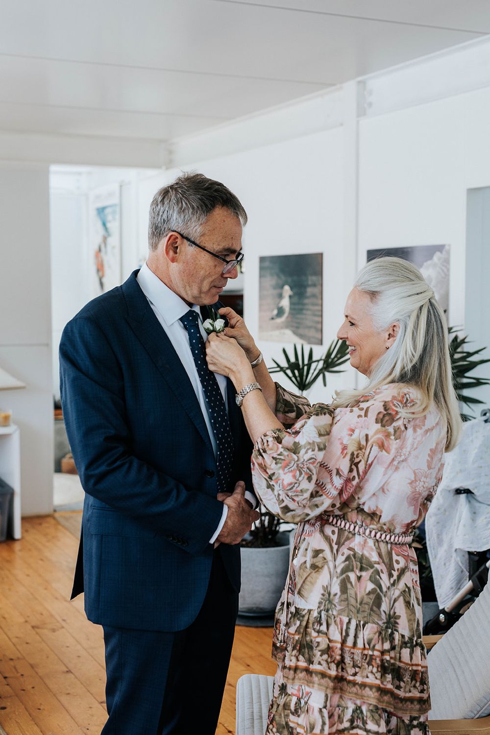 A woman in a floral dress pins a boutonniere on a man in a navy suit at a wedding preparation.