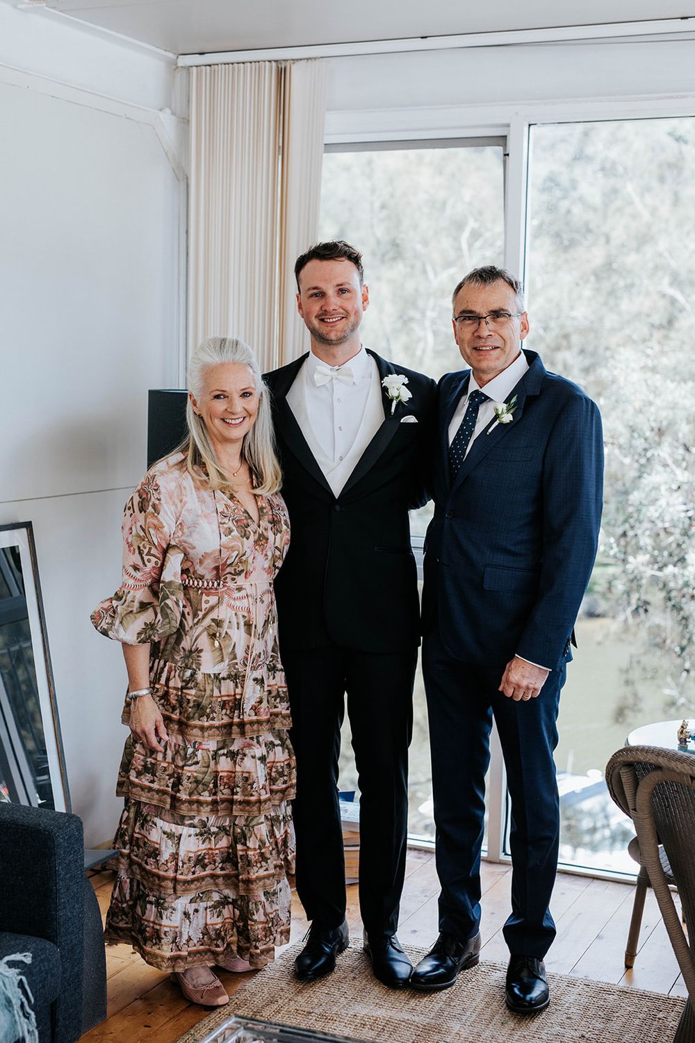 Groom in black tuxedo poses with parents, mother in floral dress and father in navy suit, indoors near large windows.