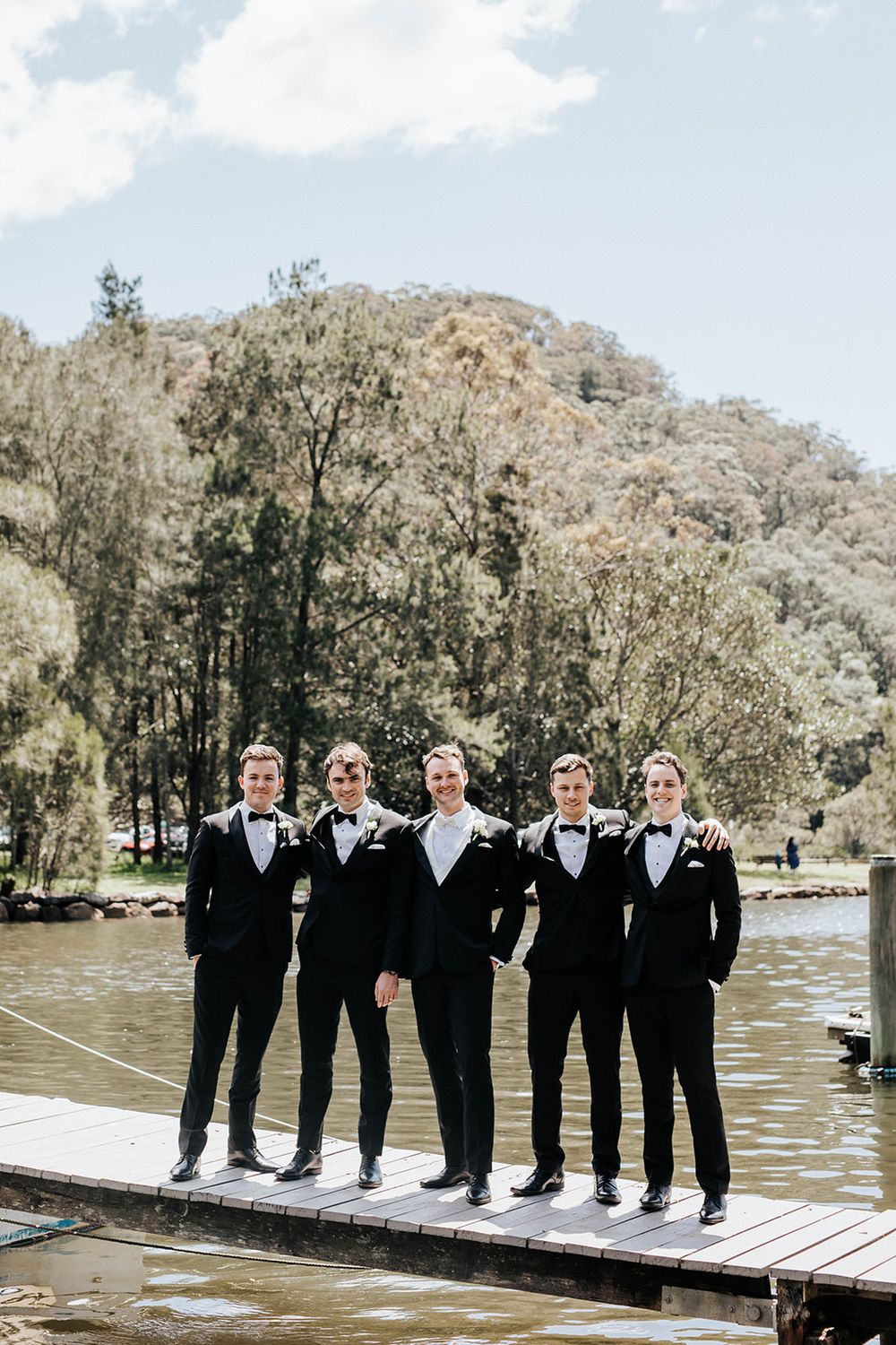 Five groomsmen in black tuxedos and bow ties posing on a wooden dock by a lake with lush green hills behind.
