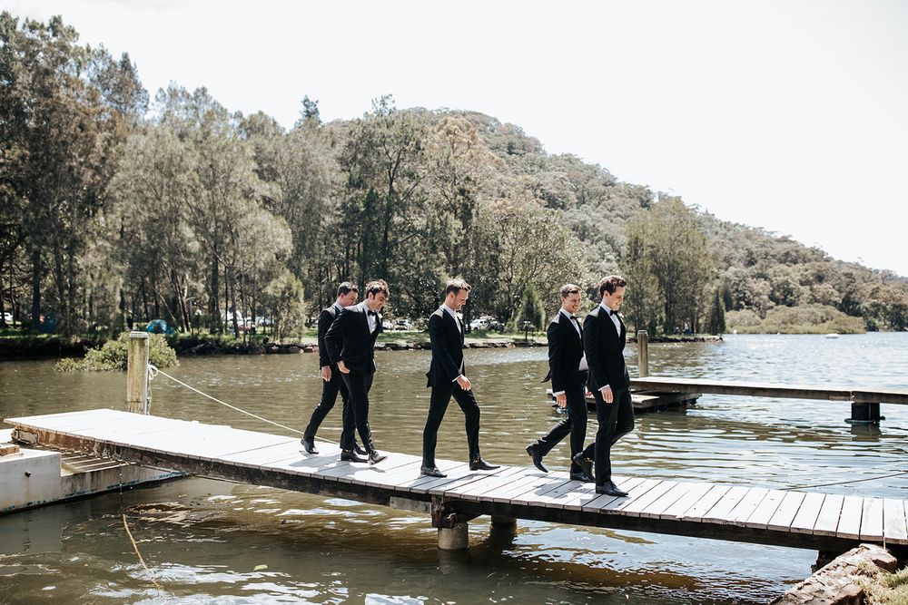 Four groomsmen in black suits walking together on a wooden dock beside a scenic lake surrounded by lush green trees.