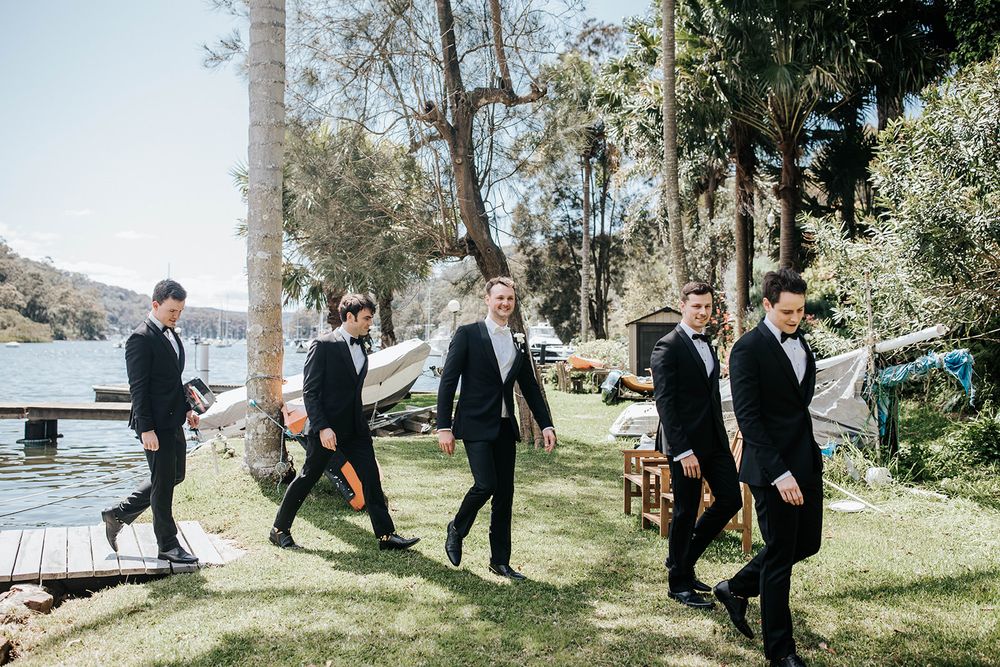 Five groomsmen in black suits walking together on green lawn near waterfront trees on a sunny wedding day.