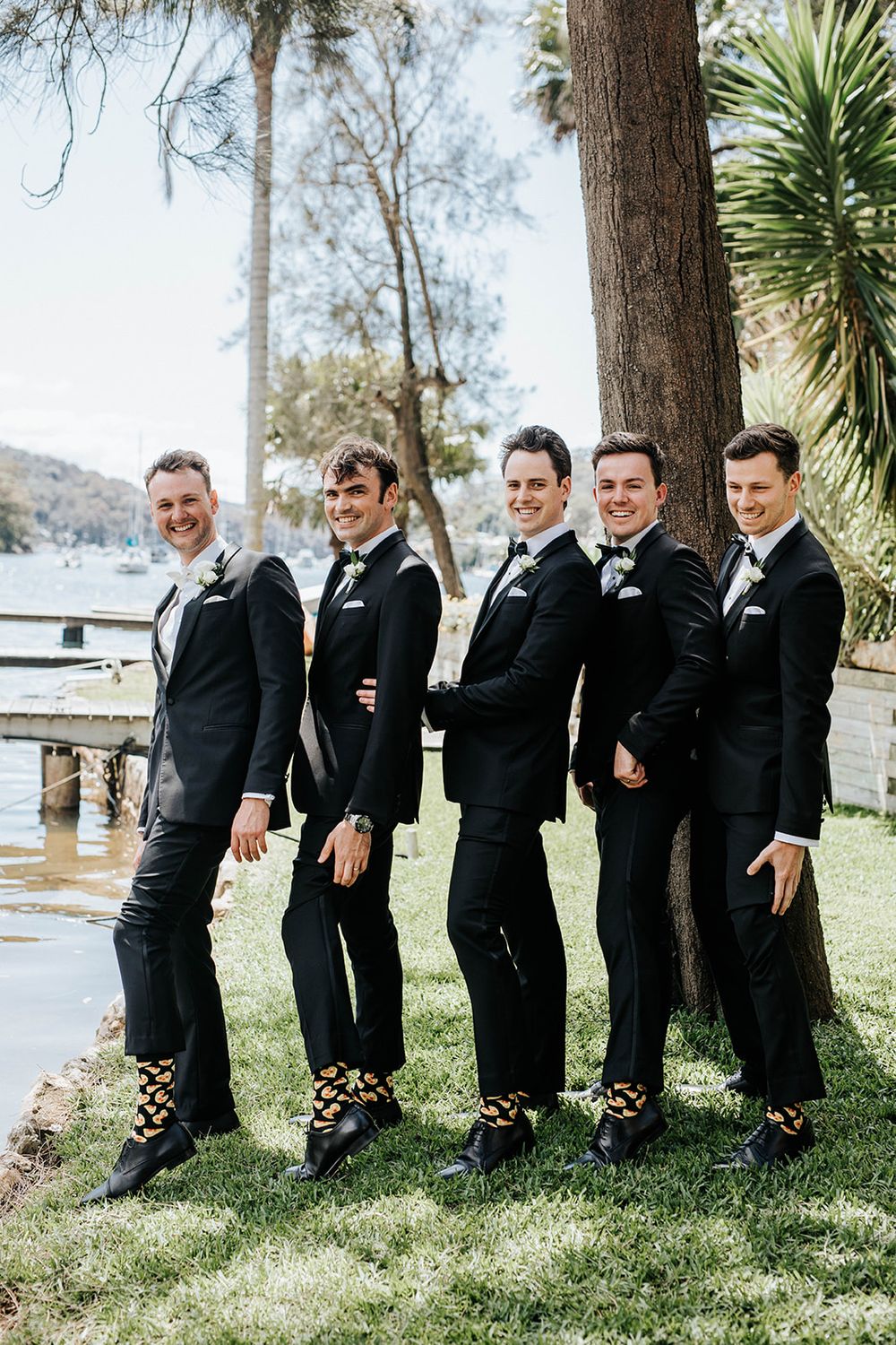 Five groomsmen in black tuxedos and fun patterned socks pose outdoors near a waterfront on a wedding day.