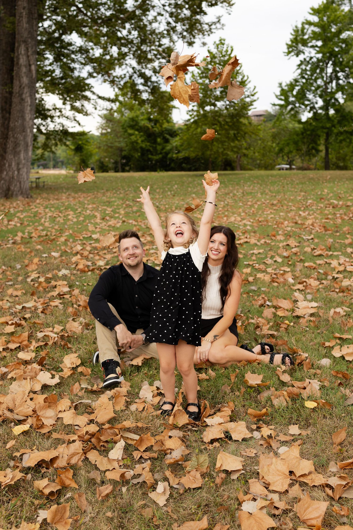 Family of three sitting in autumn leaves in a park, young girl joyfully tossing golden fall leaves into the air.