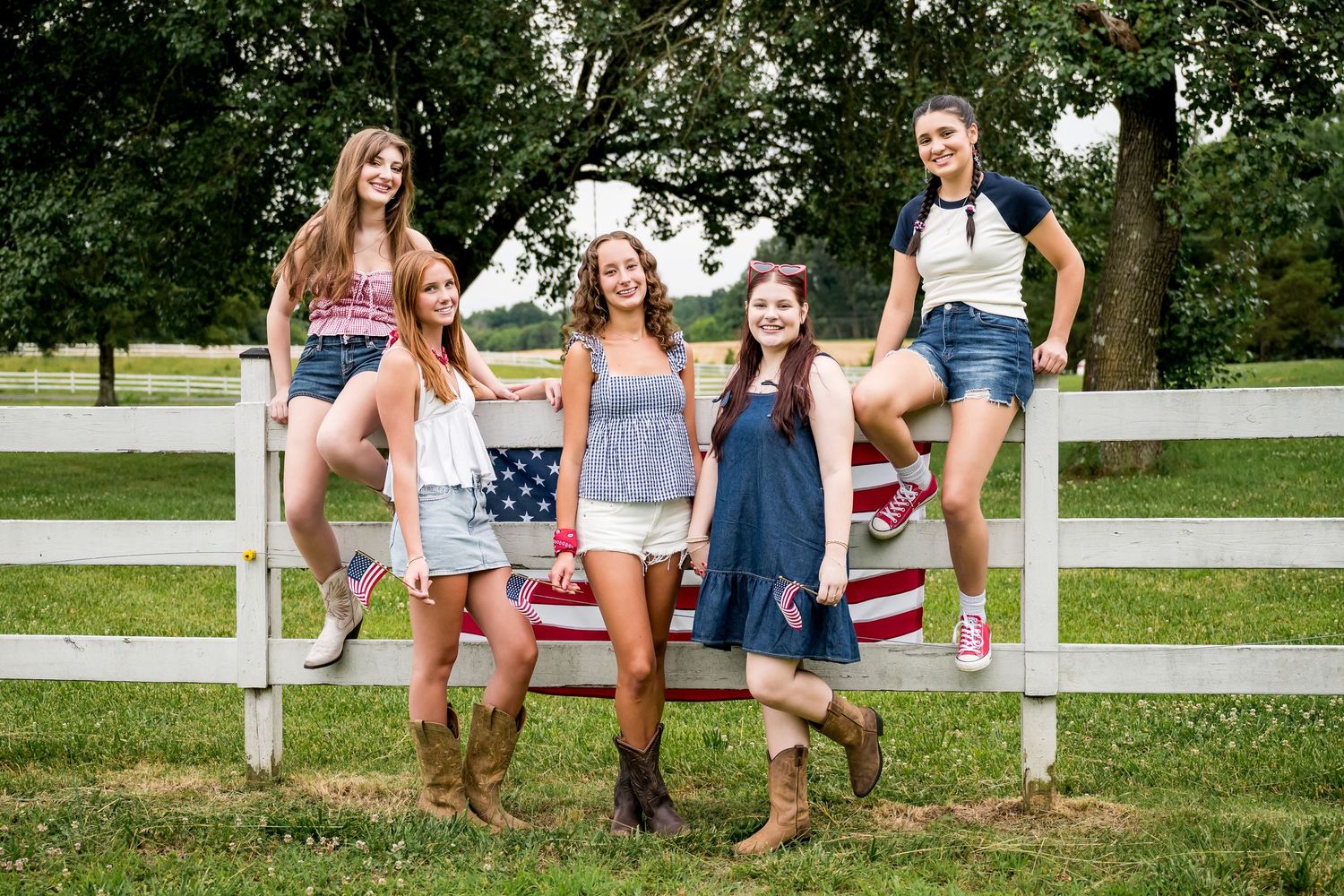 Five young women pose with an American flag on a white wooden fence in a green rural setting.