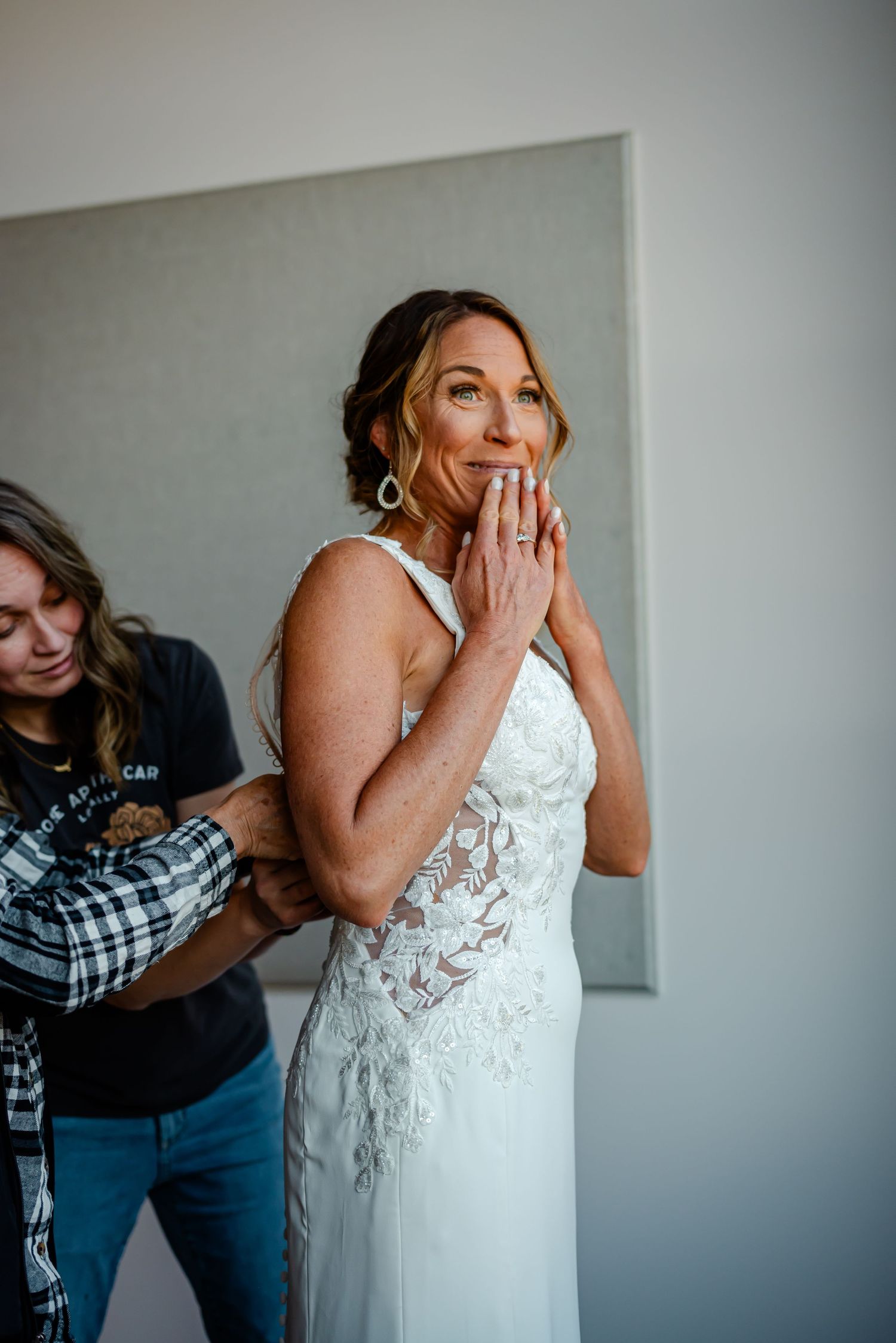 Emotional bride in lace wedding dress holds hands to face while assistant helps fasten gown before ceremony.