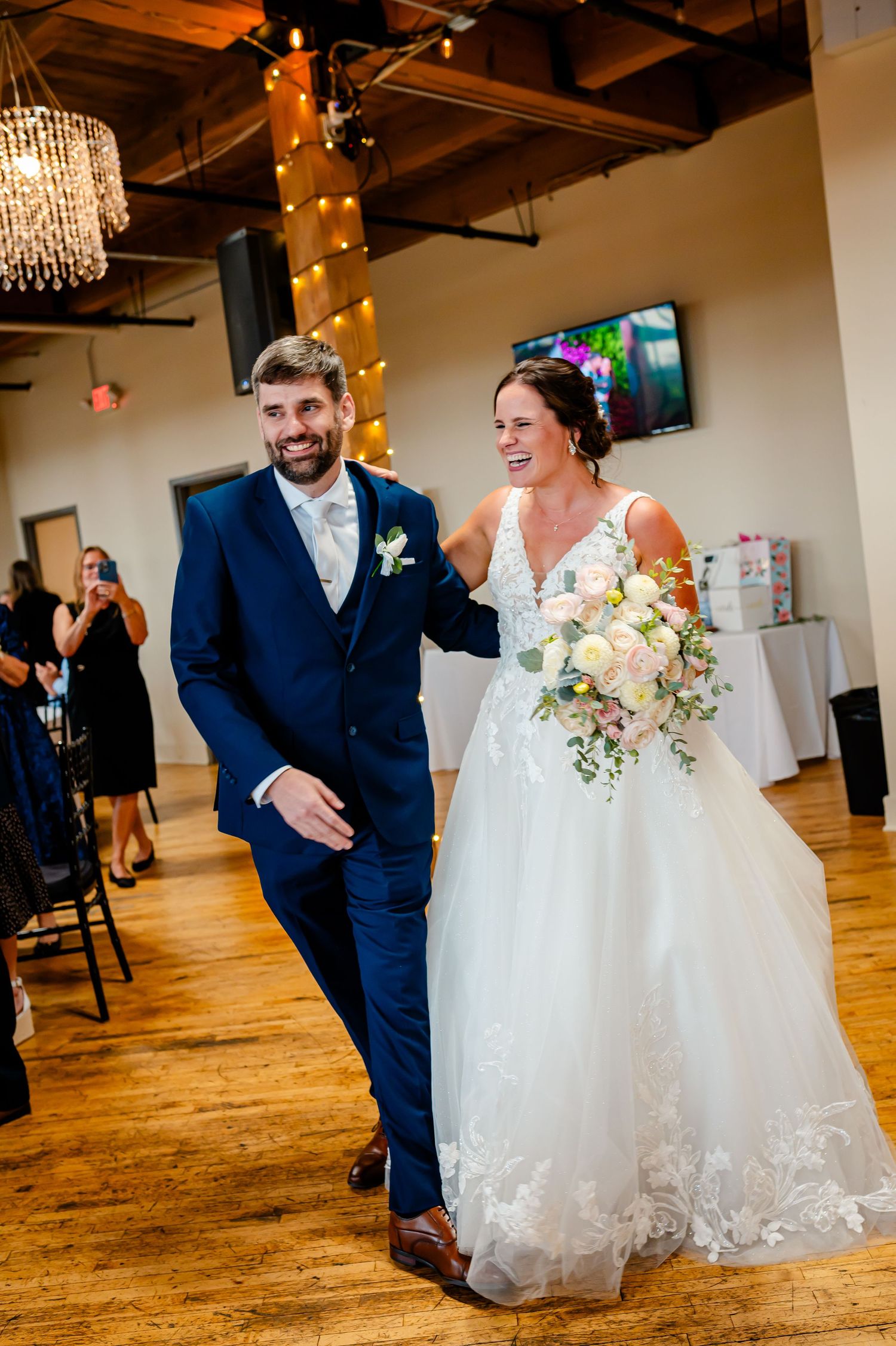 Smiling newlyweds walk together at wedding reception, bride in lace gown holding bouquet, groom in navy suit.