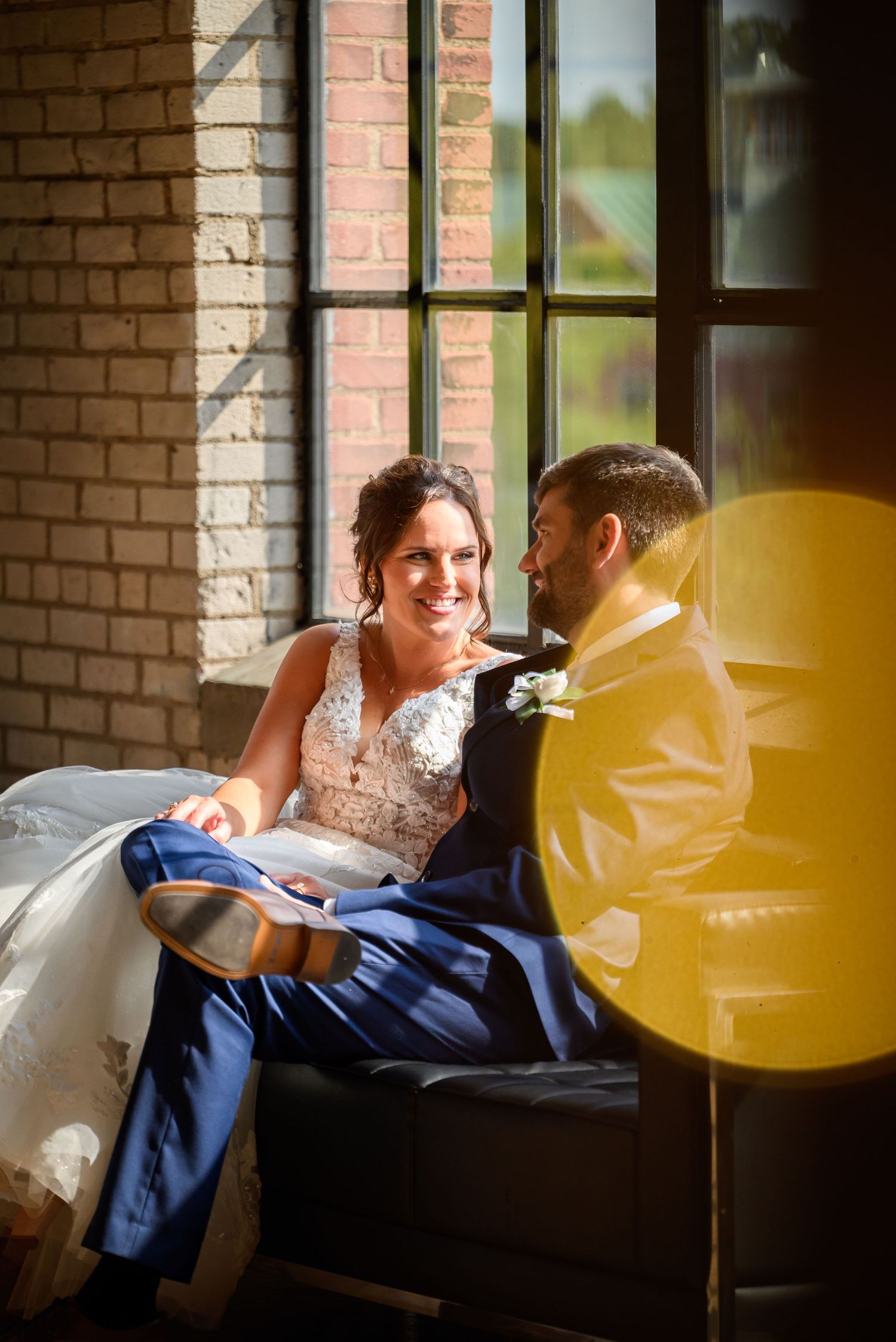 Bride and groom share a joyful moment seated by a sunlit brick window, bride showing off her wedding shoes.