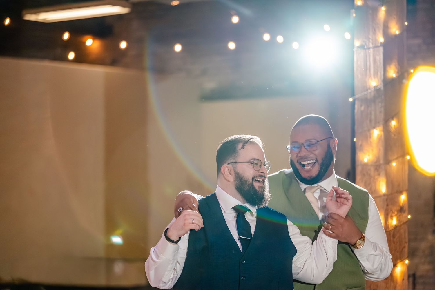 Two grooms laughing and dancing together at their wedding reception with warm string lights in the background.