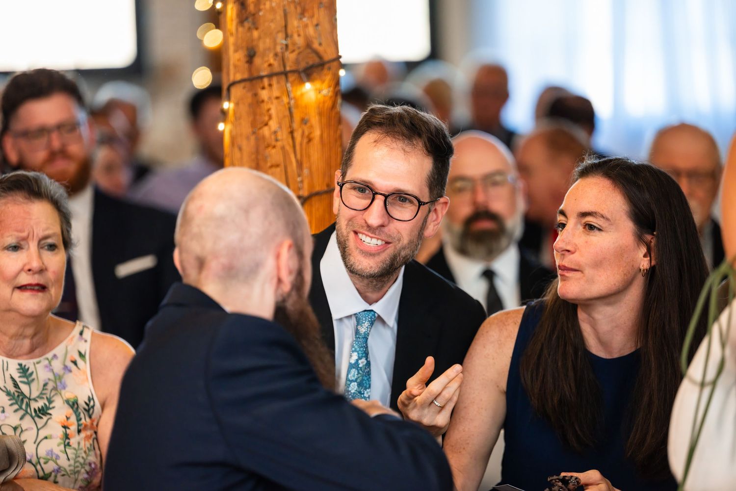 Guests socializing at an elegant wedding reception, smiling man in blue tie conversing with others in a bright venue.