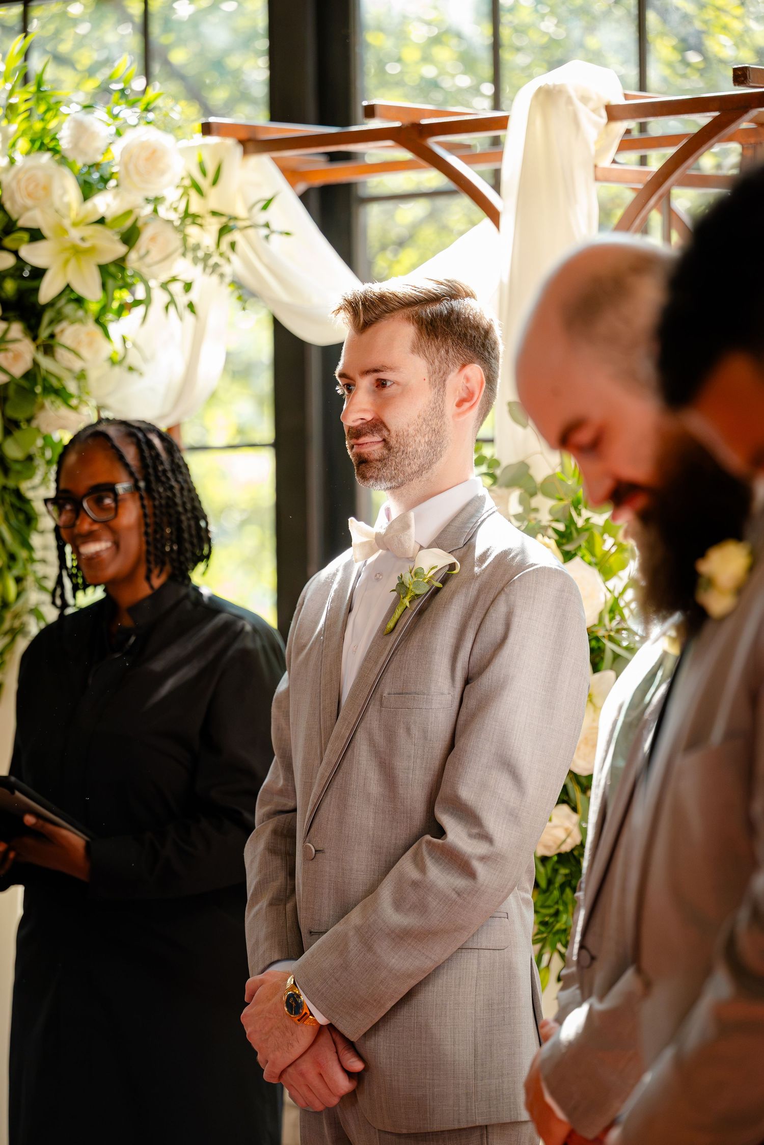 Groom in gray suit smiling at altar during wedding ceremony with floral arch and officiant.