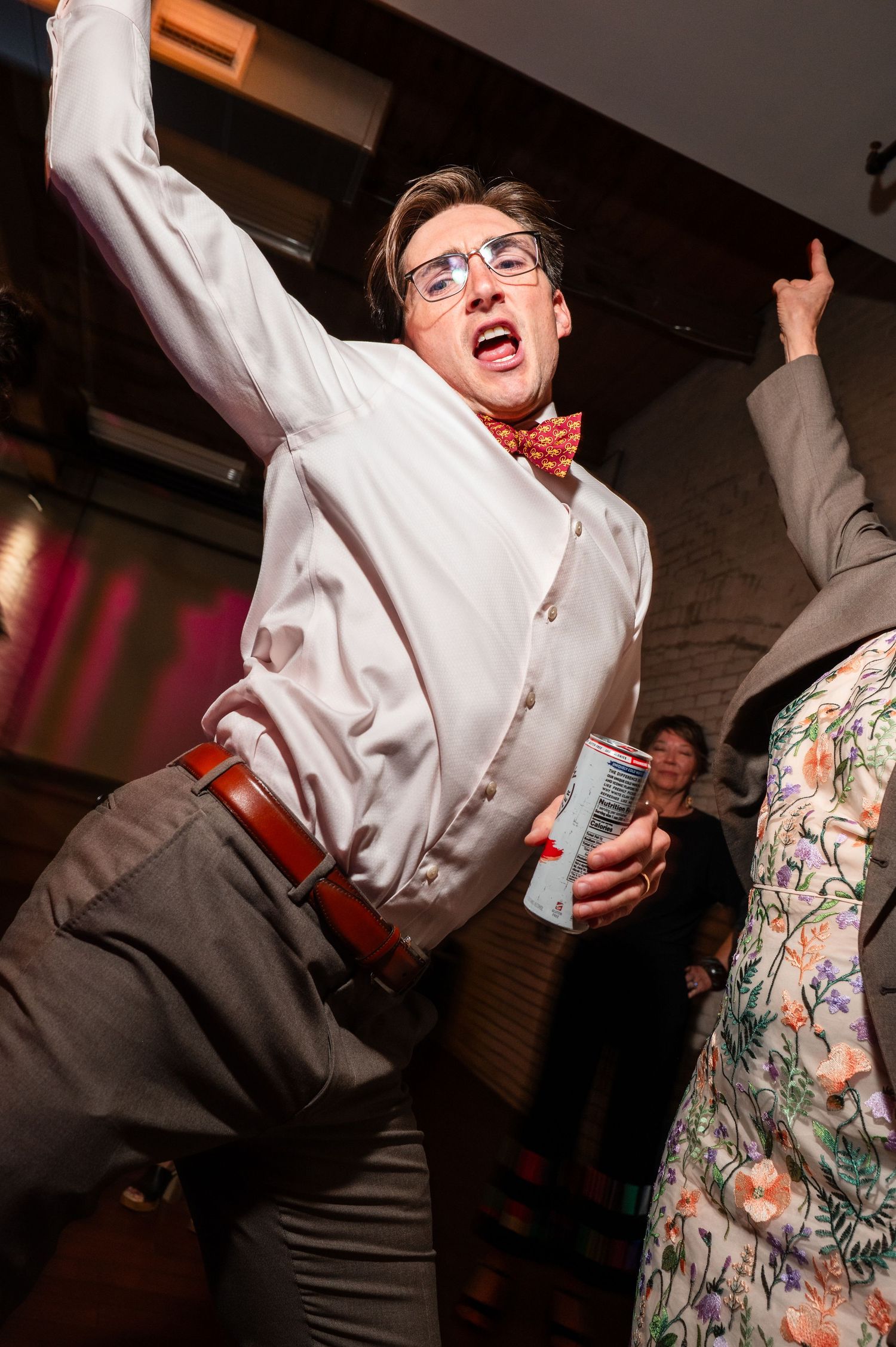 Young man in bow tie and glasses dancing energetically at a party, holding a drink can.
