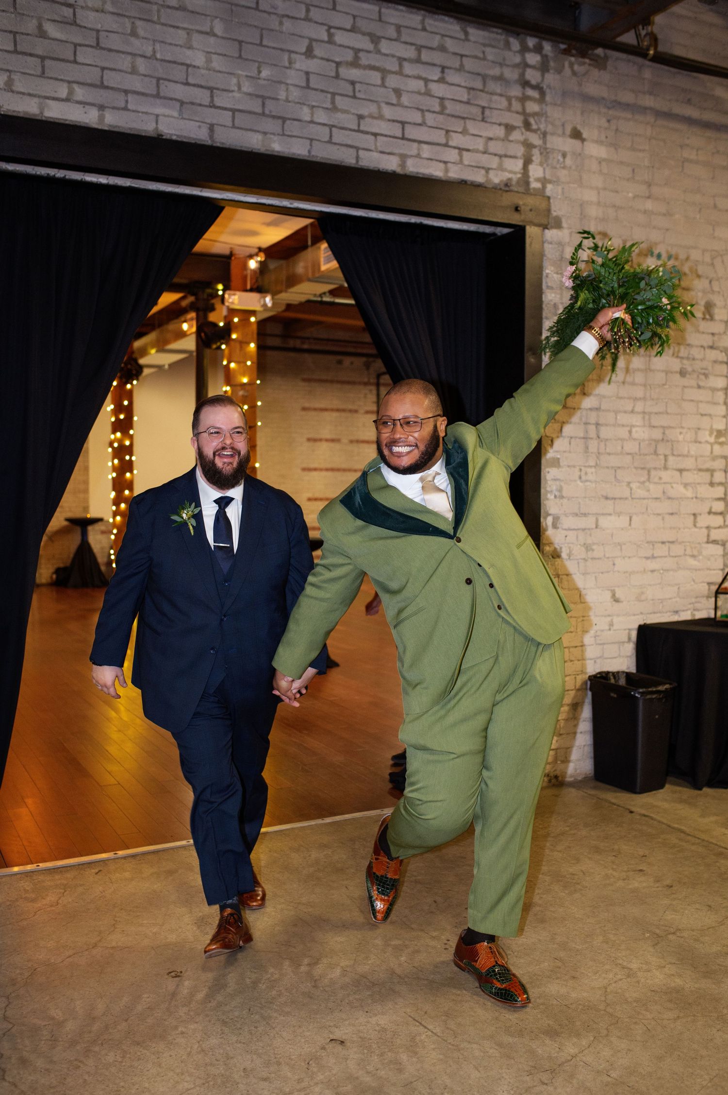 Two grooms celebrate their wedding entrance, one in green suit raising bouquet, other in navy suit, holding hands.