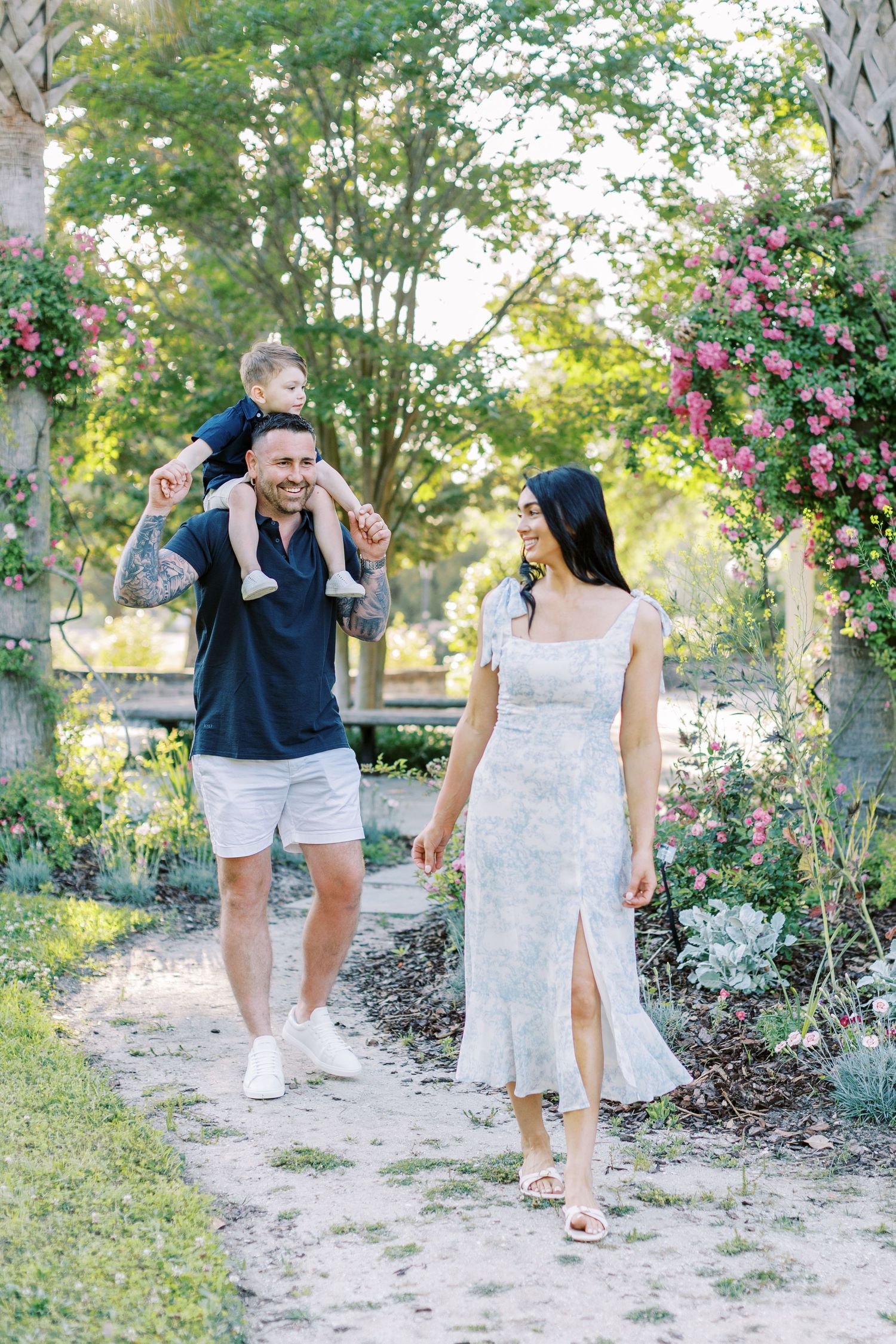 A happy family walks through a blooming garden, with a man carrying a toddler on his shoulders beside a woman in a floral dress.
