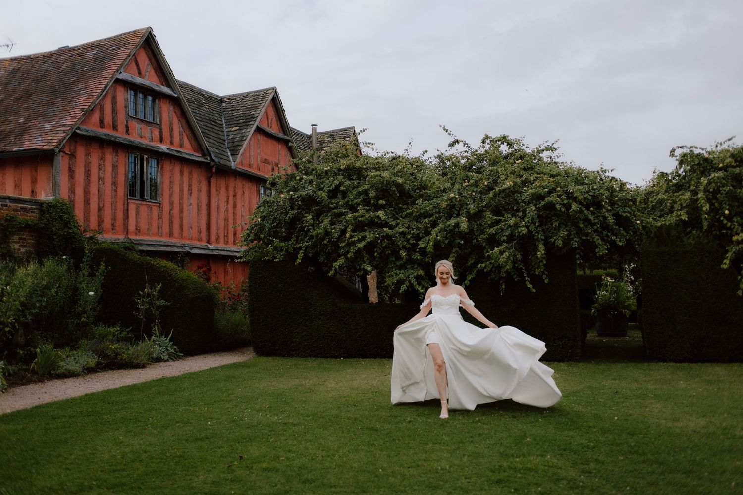 Bride in flowing white gown twirling on green lawn before a historic Tudor-style manor surrounded by lush hedges.