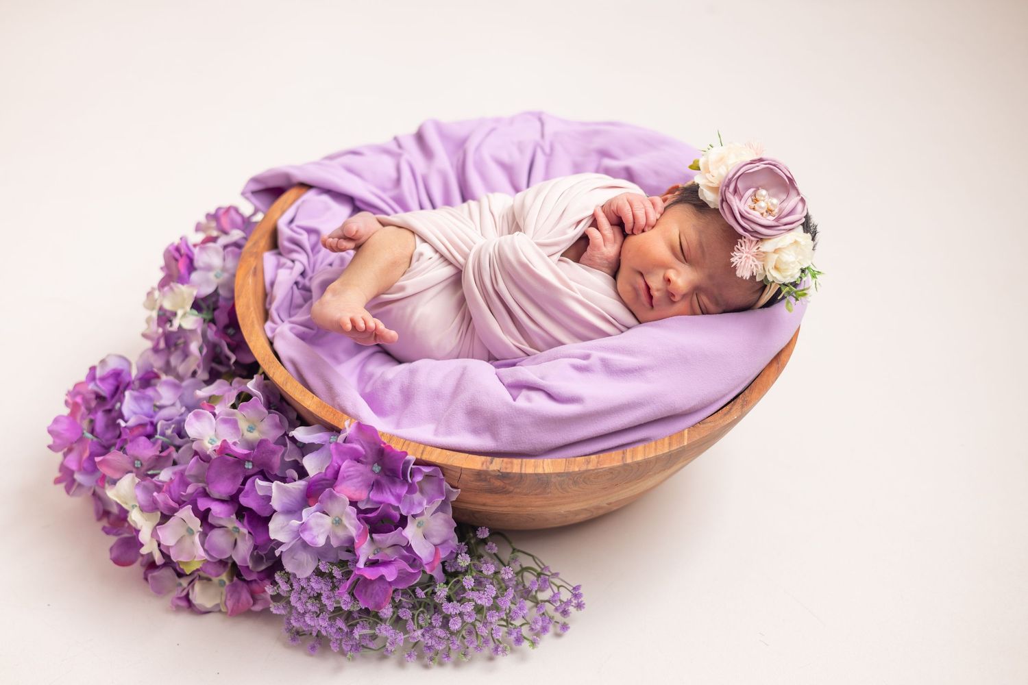 Newborn baby sleeping in wooden bowl with purple blanket and floral headband surrounded by lavender hydrangeas.