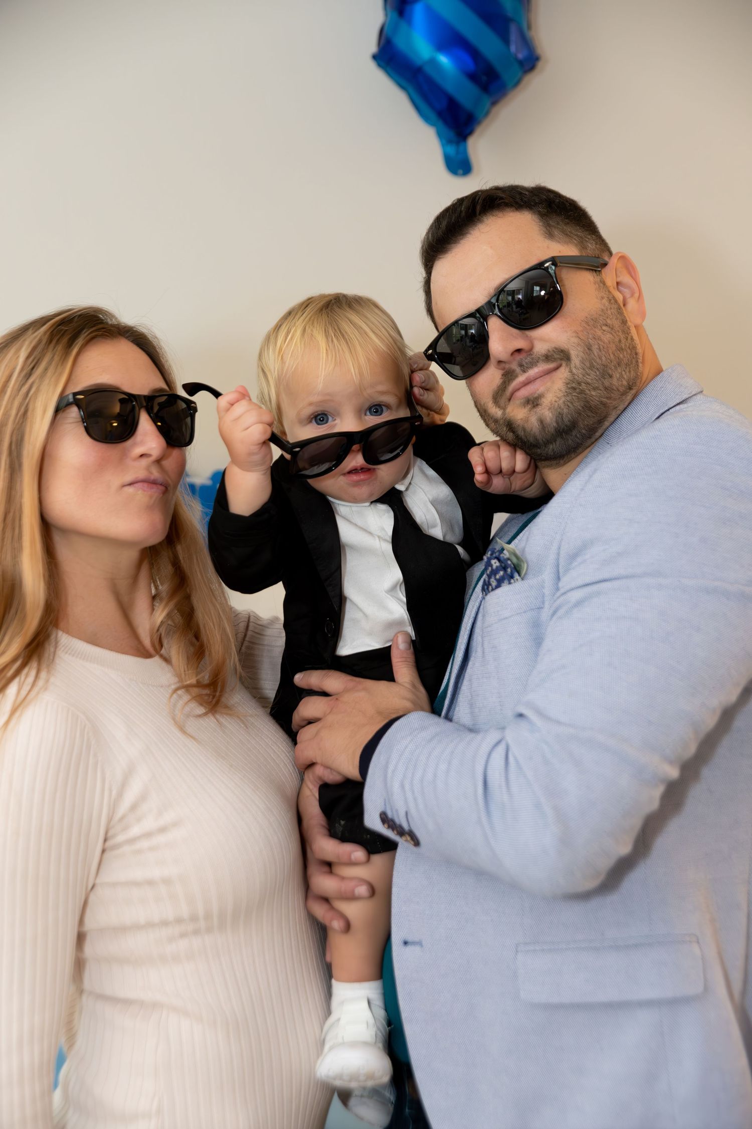 Family of three wearing sunglasses, parents holding baby in suit at birthday party with blue balloon.