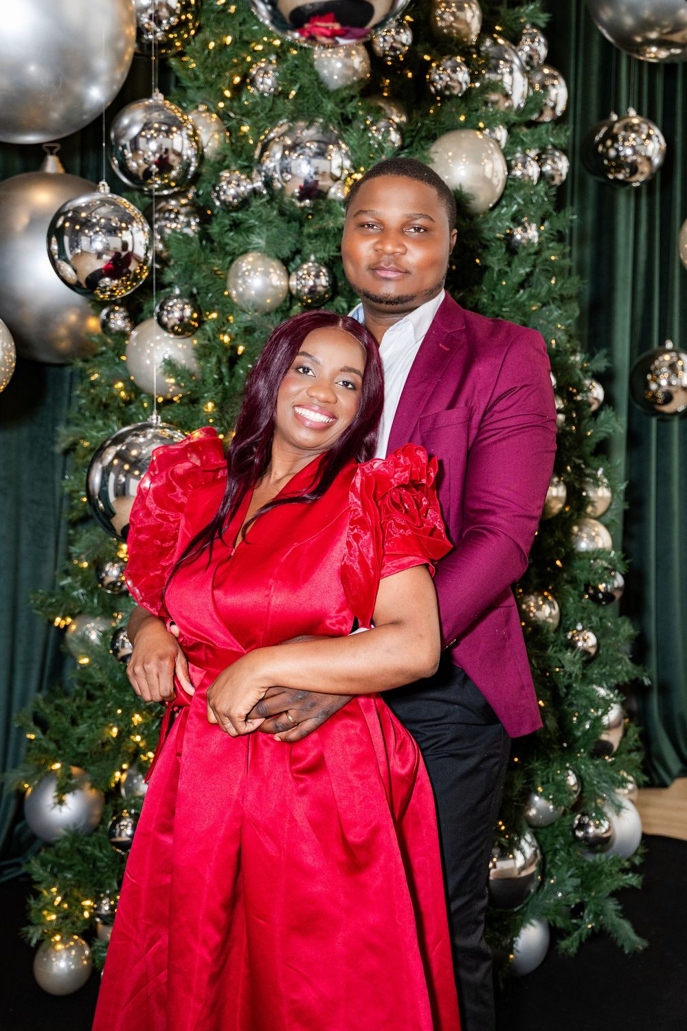 A couple in festive attire poses before a decorated Christmas tree with silver ornaments.