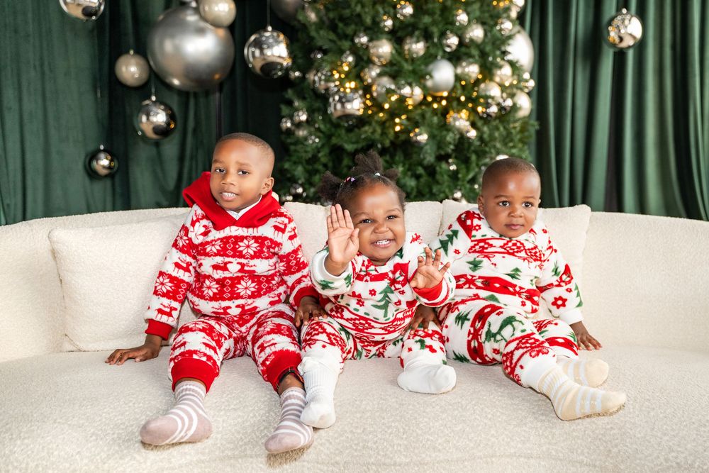 Three young children in matching Christmas pajamas sitting on a couch in front of a decorated holiday tree.