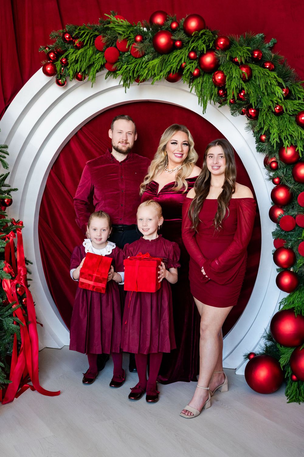 Family of five posing in festive red outfits inside a circular arch decorated with Christmas ornaments and greenery.