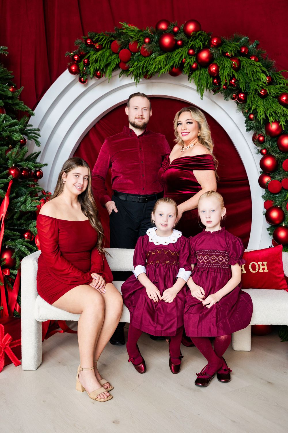Family of five posing for a festive Christmas portrait in coordinating burgundy outfits with holiday greenery and red ornaments.